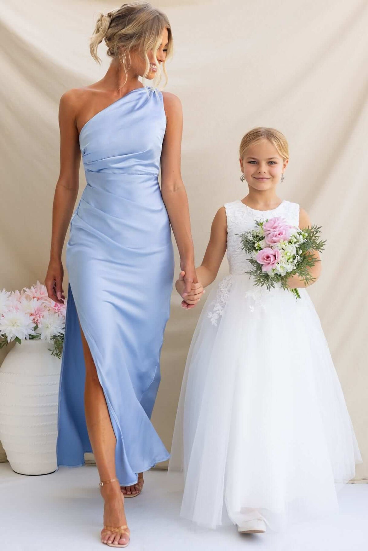 Bride in a blue one-shoulder maxi dress holding hands with a flower girl in a white dress, both smiling.