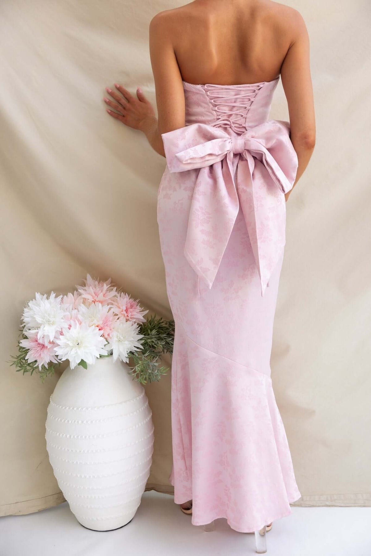 Back view of strapless pink Juanita Maxi Dress with lace-up tie and large bow, surrounded by flowers in a vase.