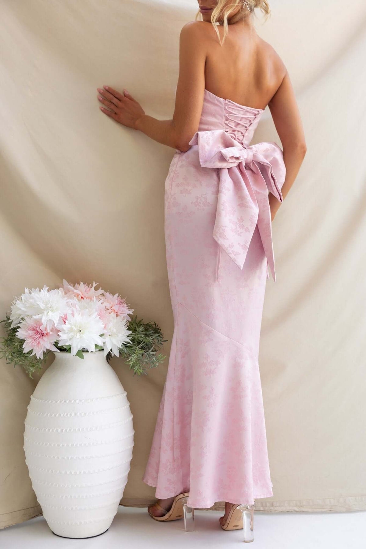 Back view of a strapless Juanita Maxi Dress in light pink, featuring lace-up detailing and a large bow.