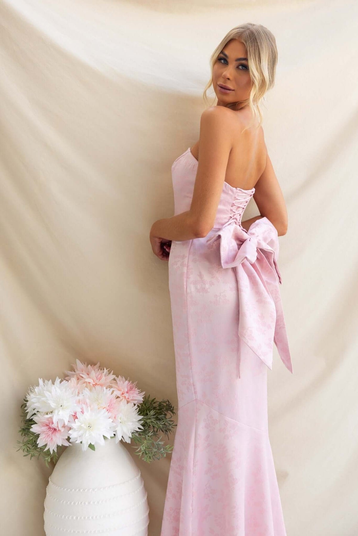 Stylish woman in a strapless pink Juanita Maxi Dress with a large bow at the back, surrounded by floral decor.