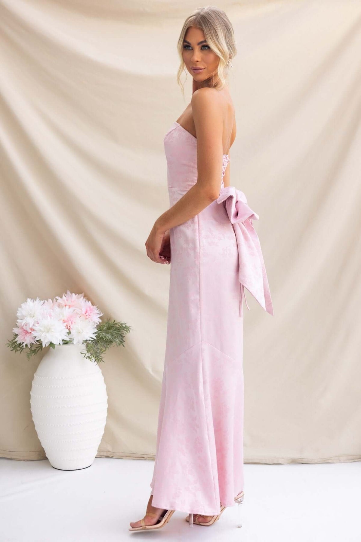 Juanita Maxi Dress in soft pink color, featuring a strapless design and a large bow at the back, styled elegantly.