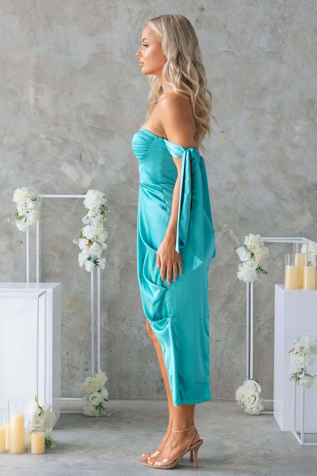 Qandice Midi Dress in teal satin with tie straps, padded bust, and asymmetrical hem, showcased on model against a floral backdrop.