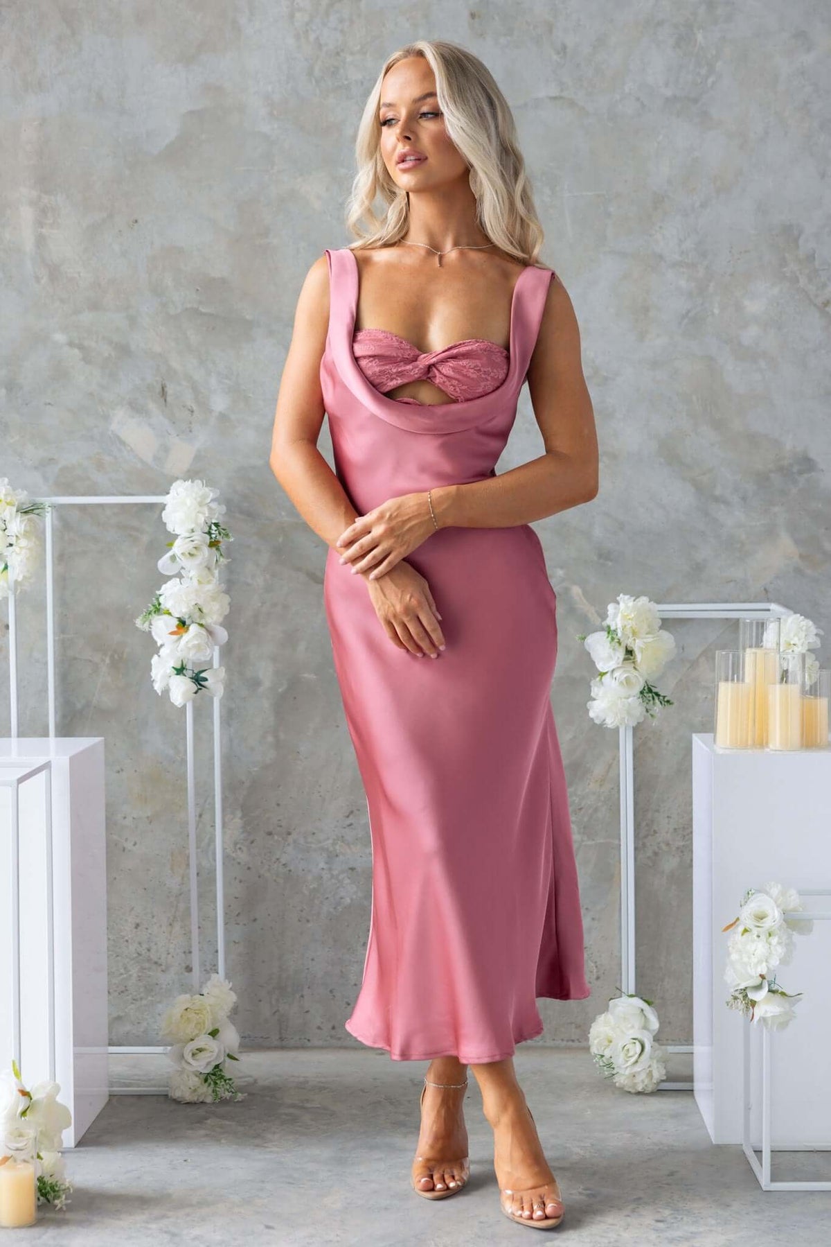 Model wearing a pink satin midi dress with scoop neckline and lace panelling, styled elegantly in a floral setting.