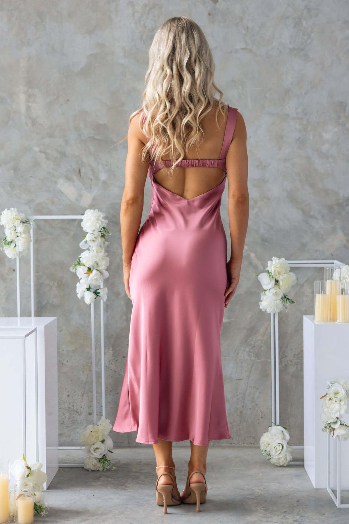 Back view of a model in a pink midi dress with a scoop neckline and lace detailing, styled with a soft satin finish.