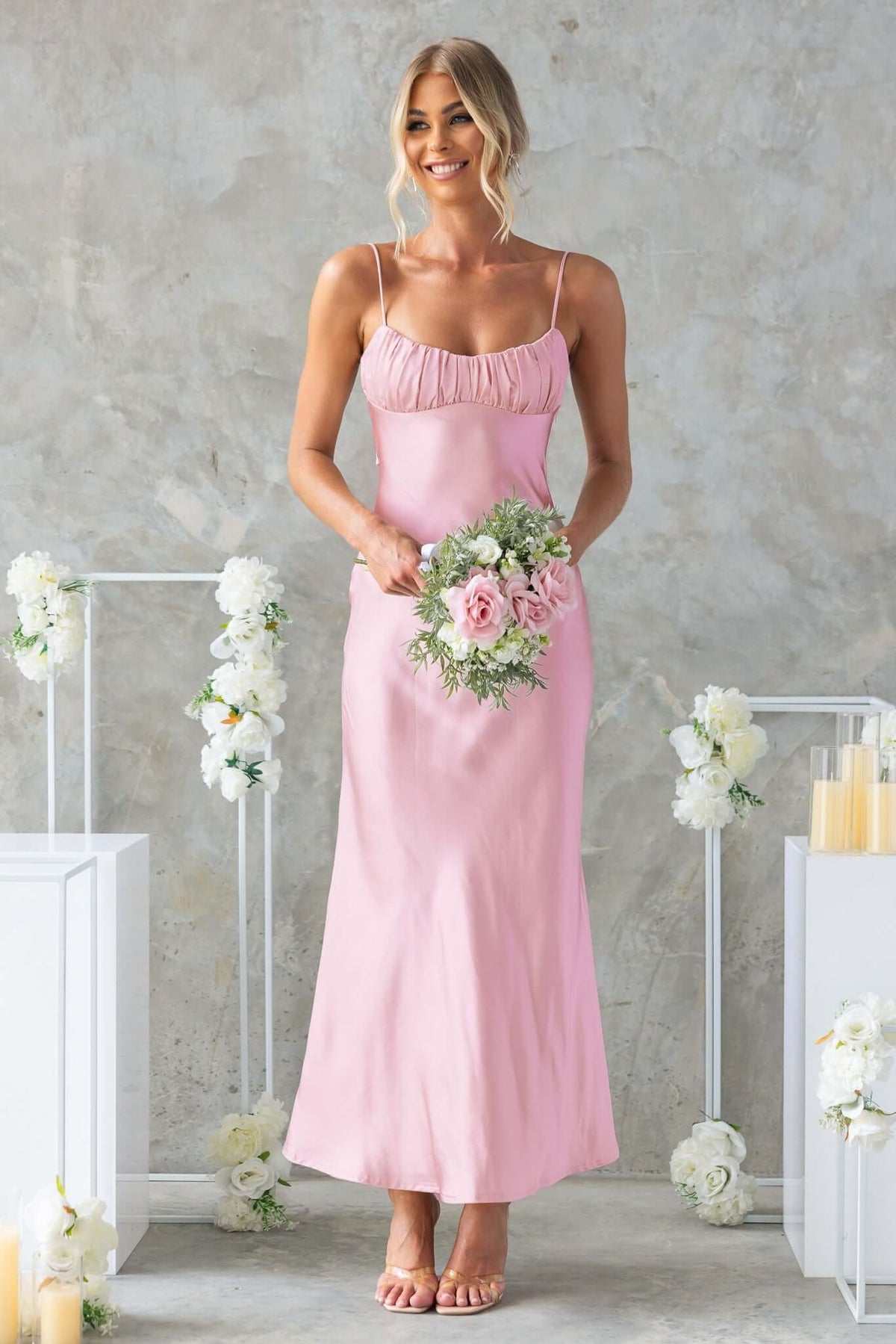 Model wearing a pink Maeya Maxi Dress with adjustable straps, holding a bouquet in a floral setting.