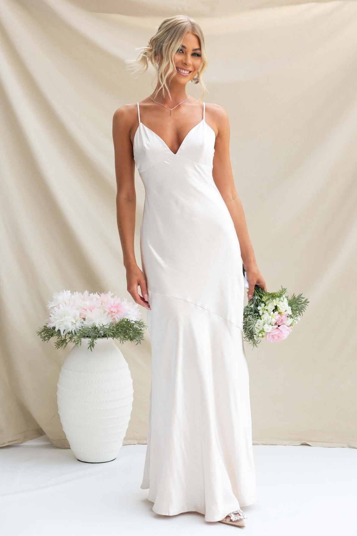 Model wears the Coralina Maxi Dress, featuring adjustable straps and satin fabric, holding a bouquet in a light setting.