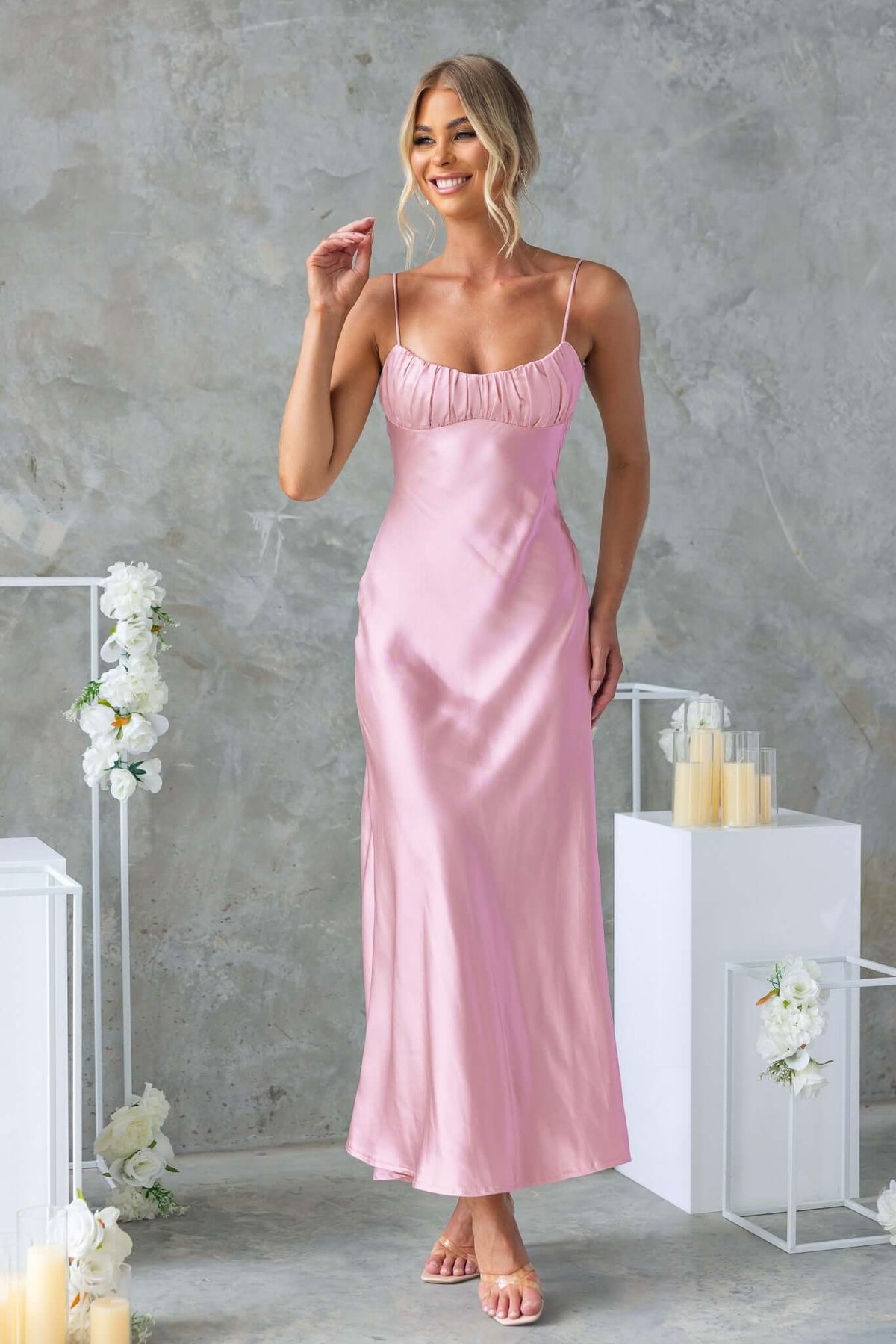 Model wearing Maeya Maxi Dress in soft pink satin with adjustable straps and gathered bust design, set against a floral backdrop.