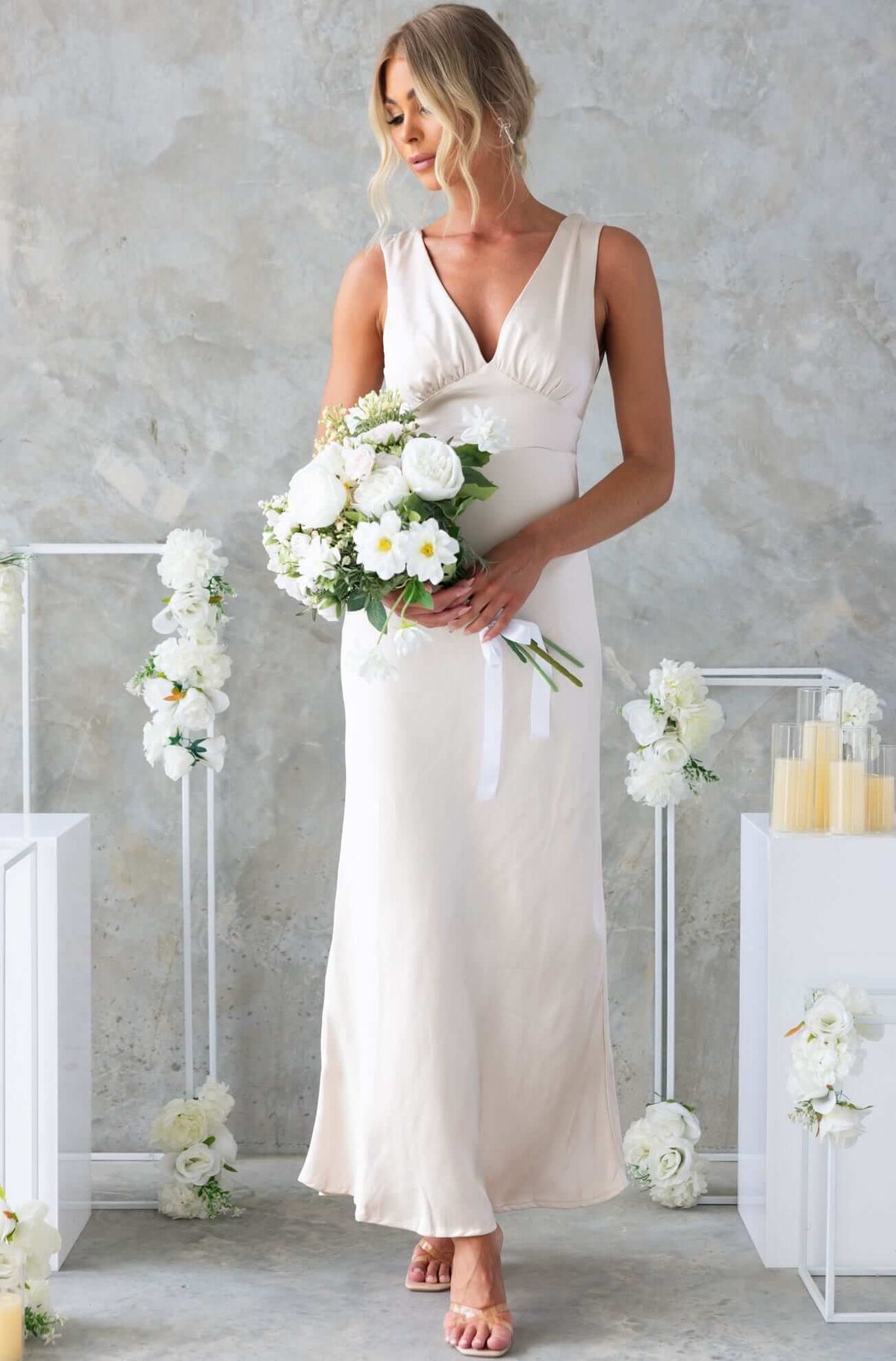 Alika Maxi Dress by Amaroso Bridal, V-neckline satin dress; model holding bouquet with floral backdrop.