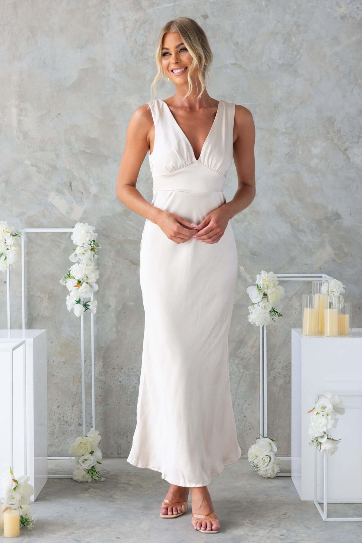Model wearing Alika Maxi Dress, a satin V-neck gown with back split and tie detail, styled elegantly among floral decor.