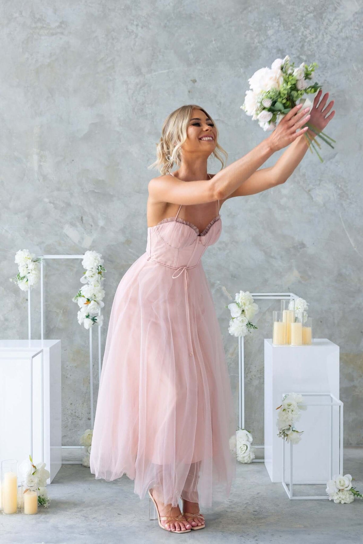 Bride in a pink tulle midi dress with adjustable straps, joyfully reaching for a bouquet against a muted backdrop.