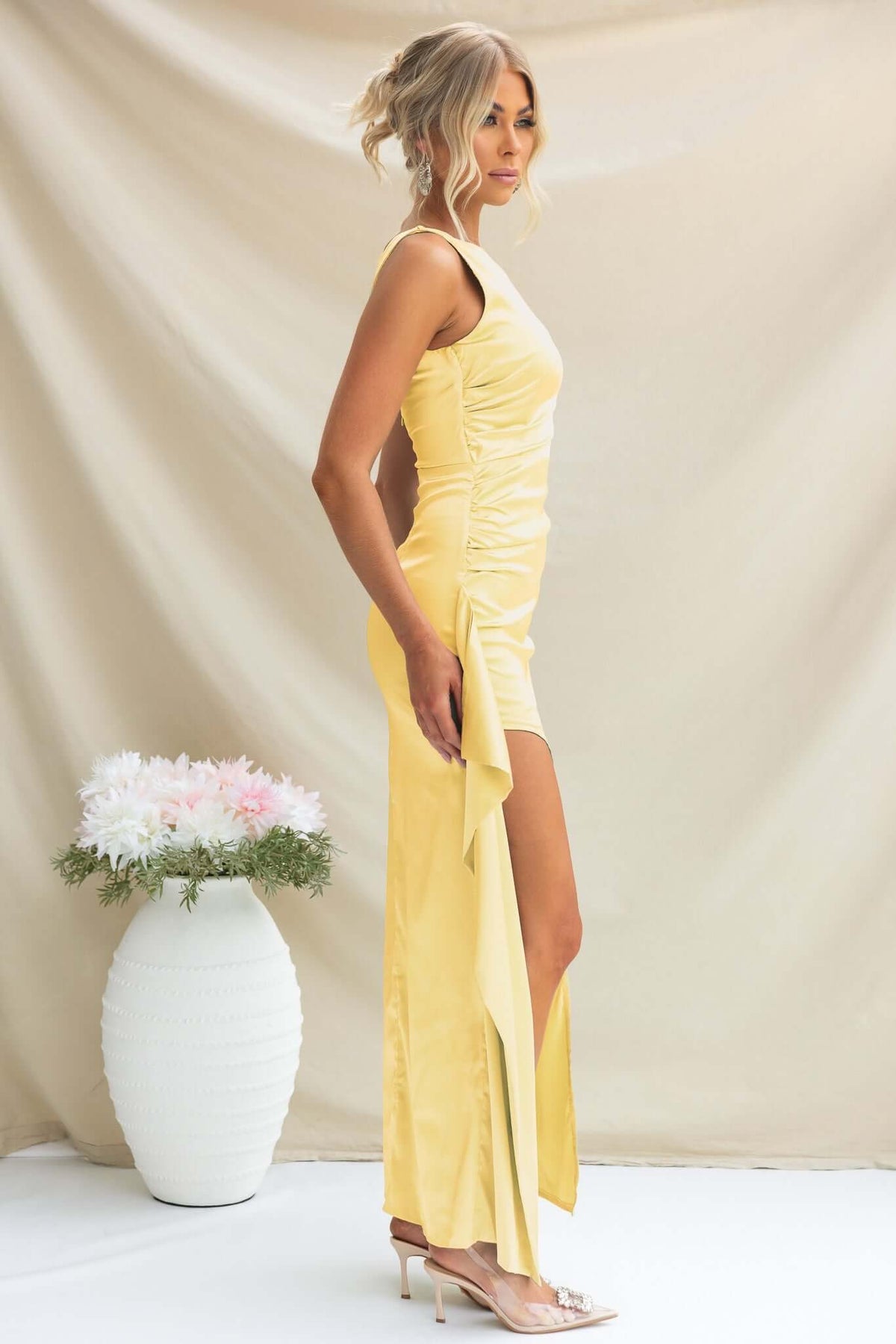 Model wearing Kenzie Maxi Dress in yellow satin with high side split and ruffle, styled with heels.