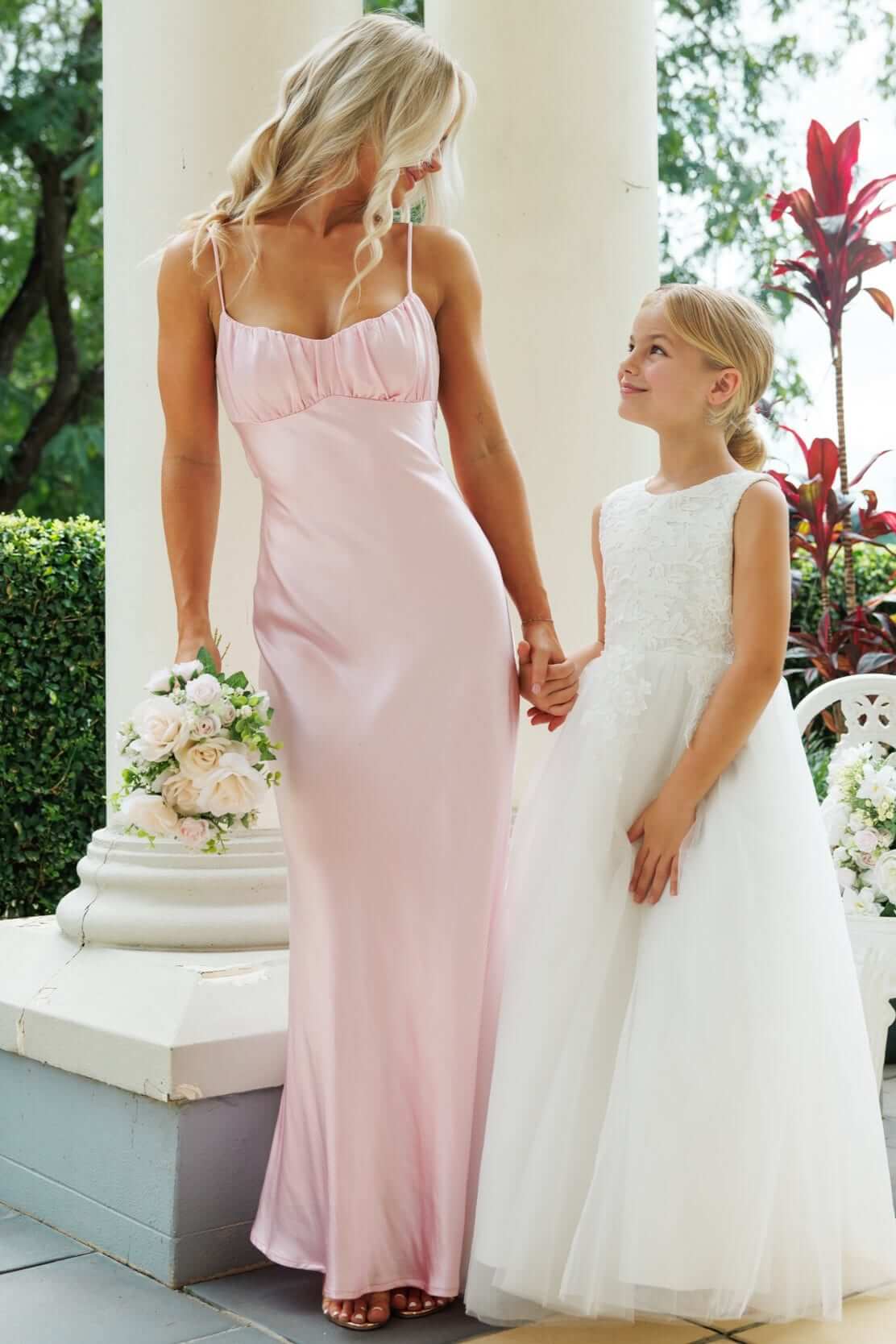 Mom and daughter in elegant dresses; adult in satin pink maxi dress and child in white flower girl gown.