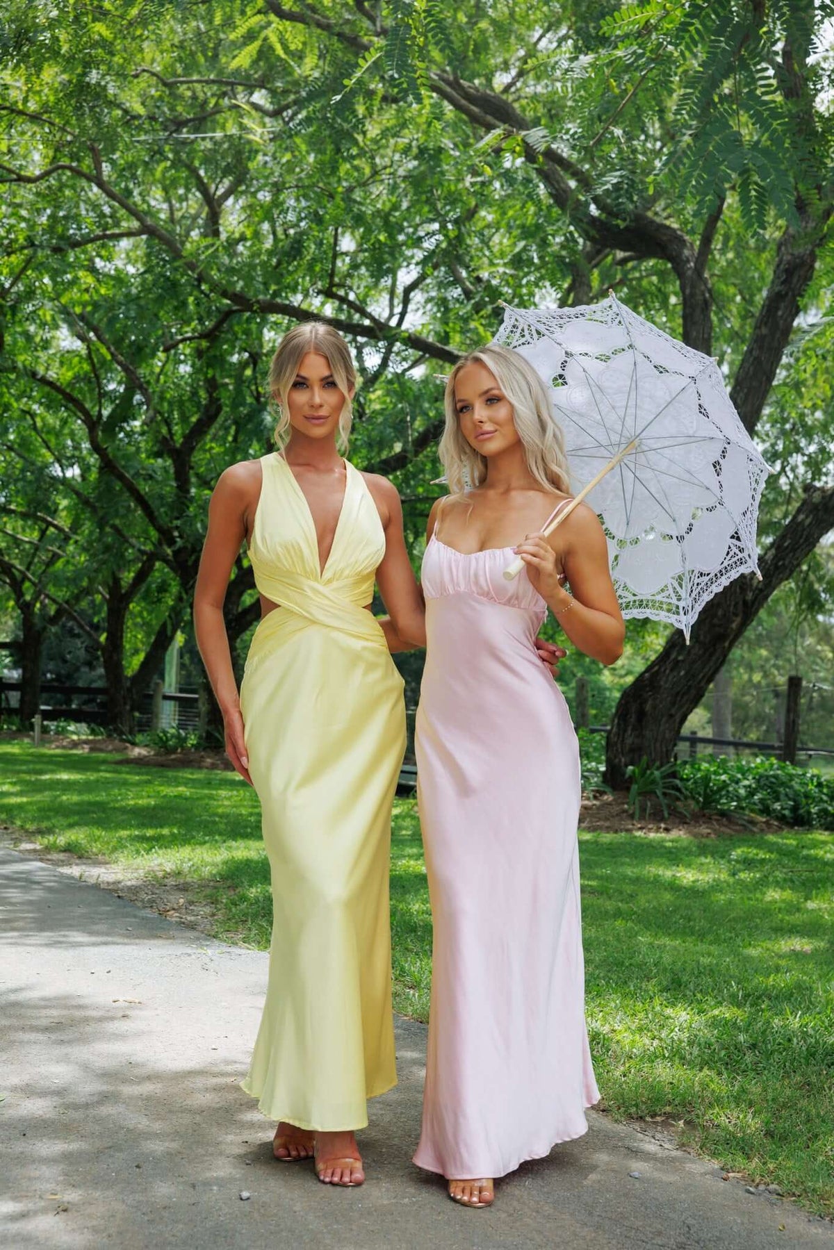 Two women in elegant maxi dresses in a sunny park, one in yellow and the other in pink, with a vintage white parasol.
