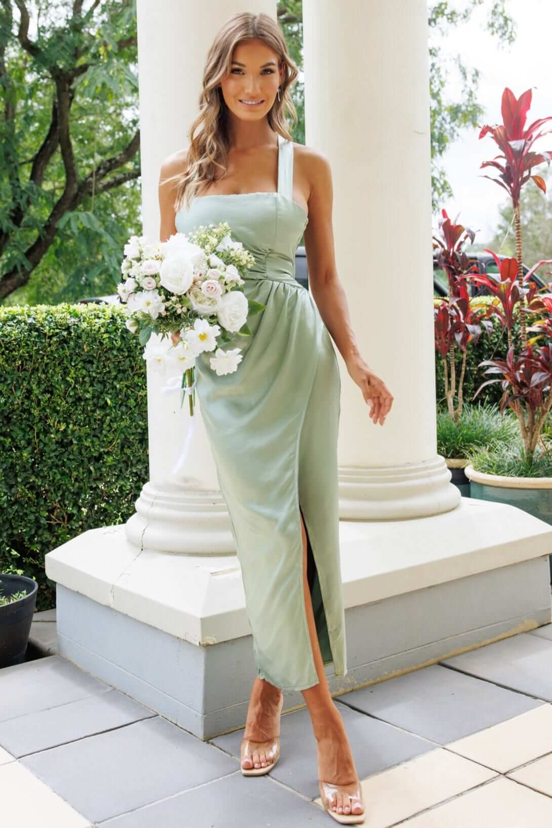 Model wearing the Merritt Maxi Dress, a green satin gown with adjustable straps and a side slit, holding a bouquet.
