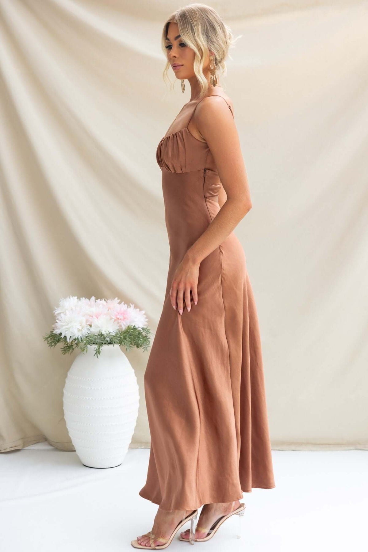 Maeya Maxi Dress in satin with adjustable straps, gathered bust, and side zip, styled with heels and flowers.