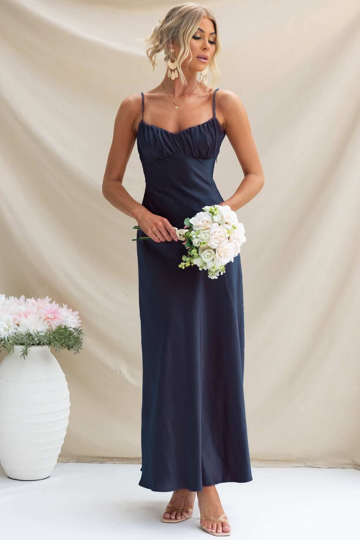 Model wearing a navy Maeya Maxi Dress with adjustable straps, holding a bouquet, against a soft background.