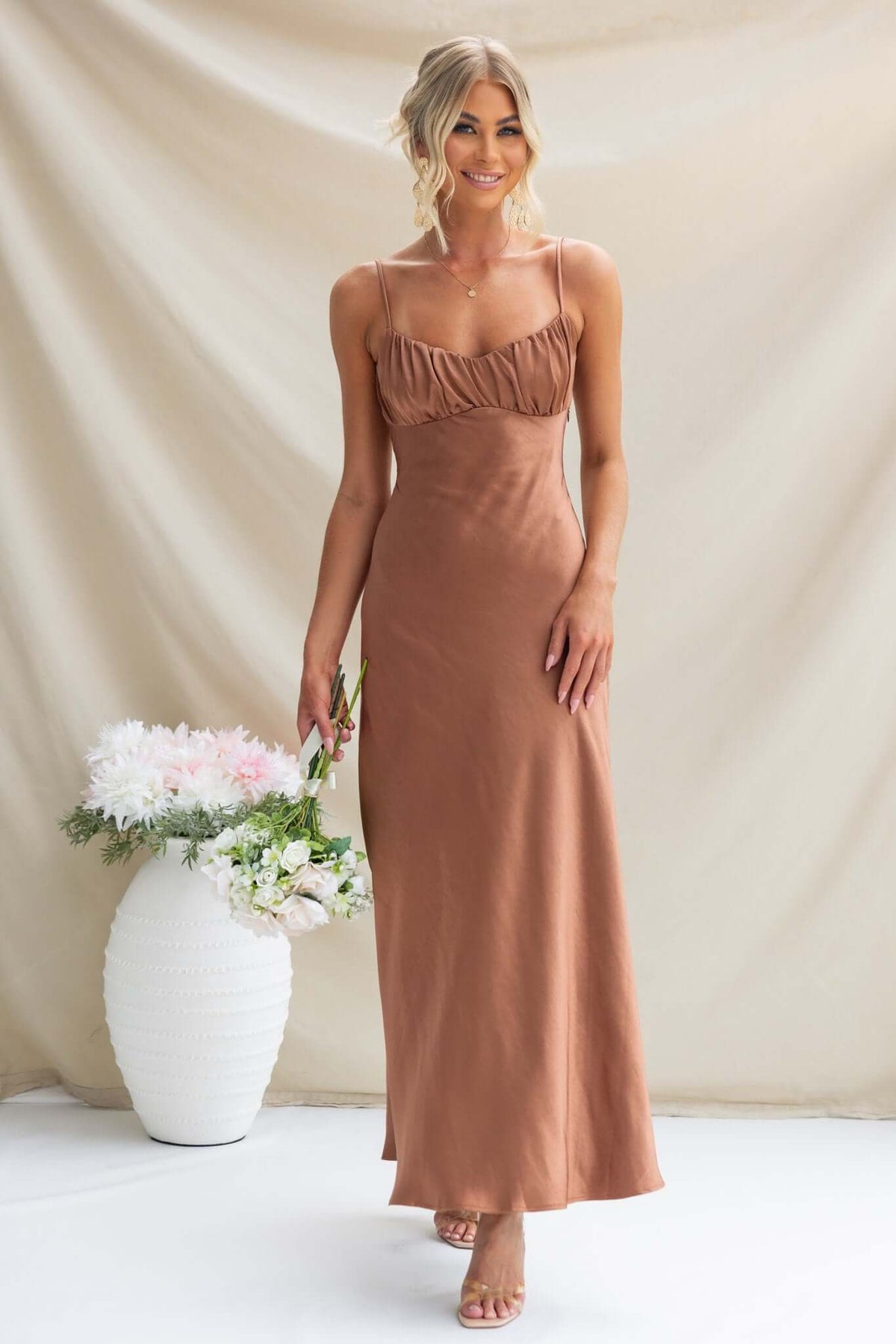 Model wearing Maeya Maxi Dress in brown with adjustable straps and gathered bust, holding flowers against a neutral backdrop.