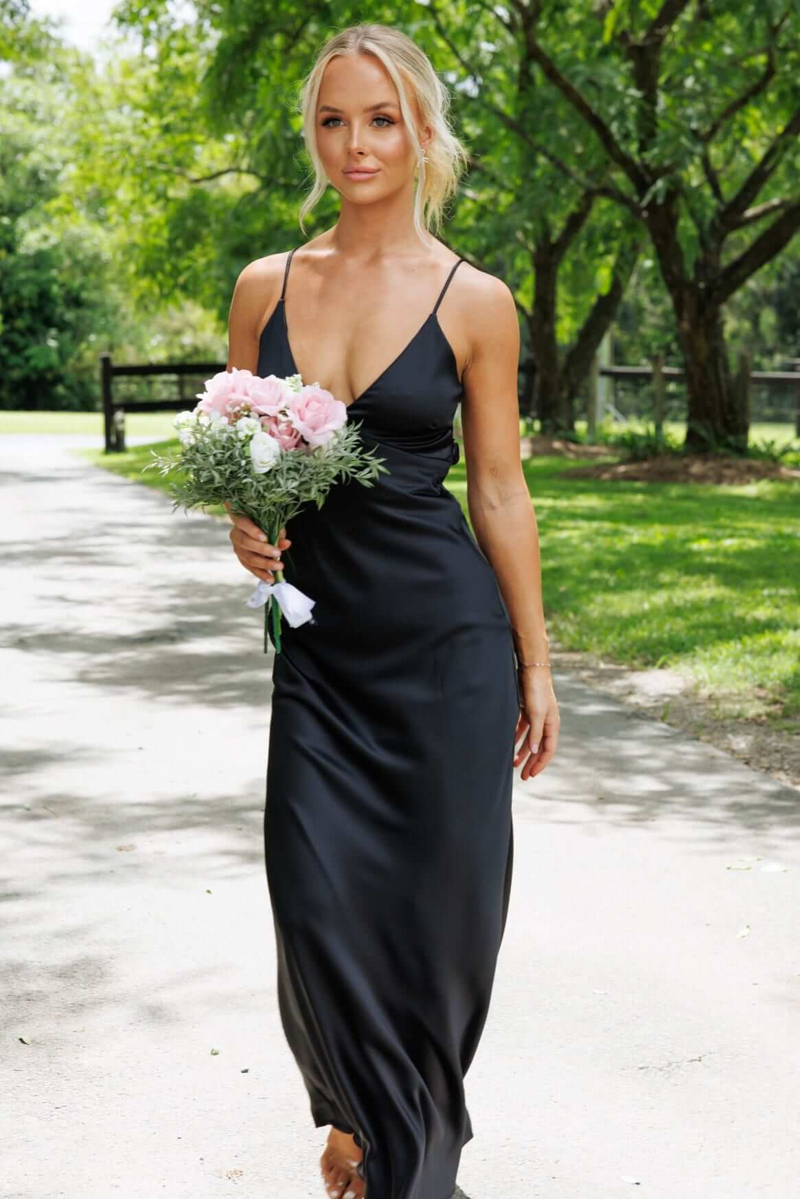 Model in black Hasina Maxi Dress with V-neckline, adjustable straps, holding bouquet in outdoor setting.
