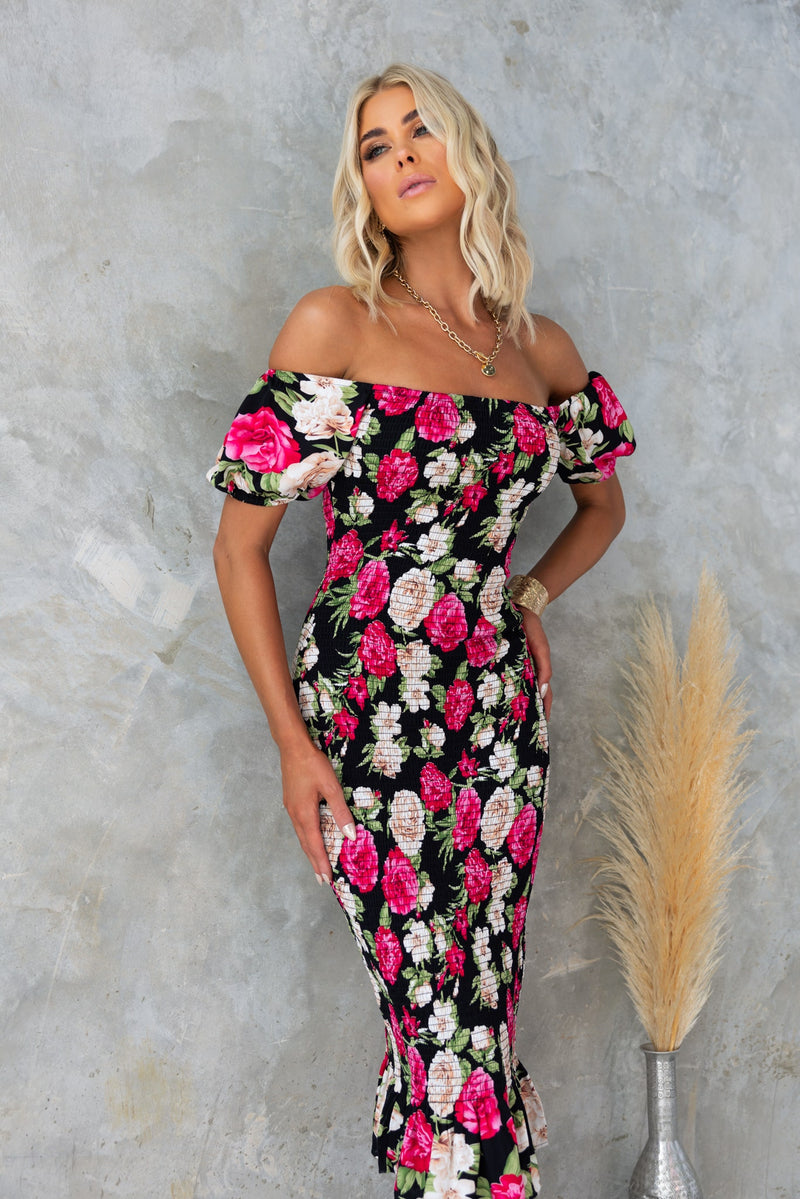 Black floral Botanic Midi Dress with off-shoulder neckline and shirred design, worn by model against a textured backdrop.
