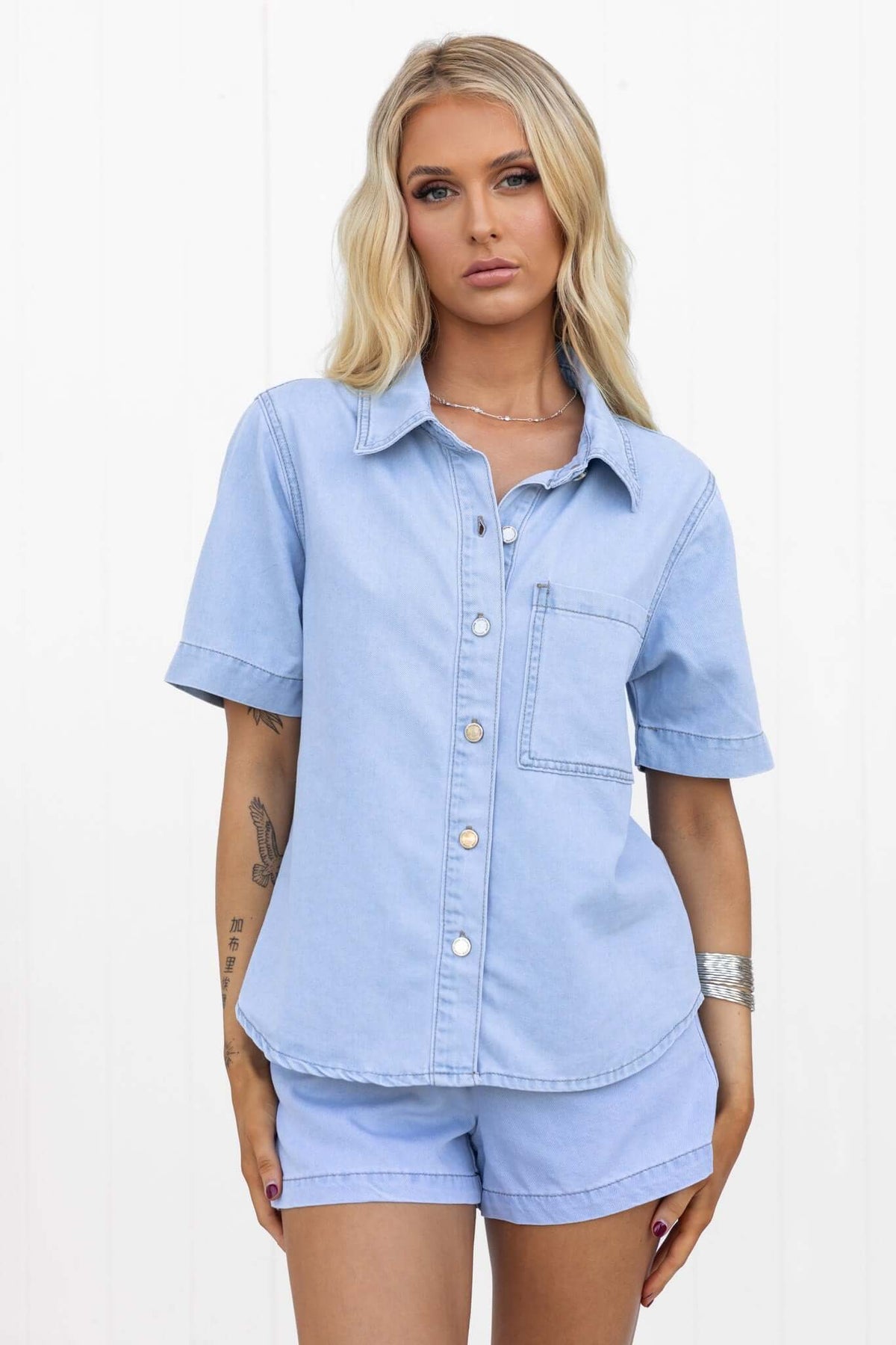Woman wearing a light chambray Winslow shirt with button-down front and pocket, styled with matching shorts.