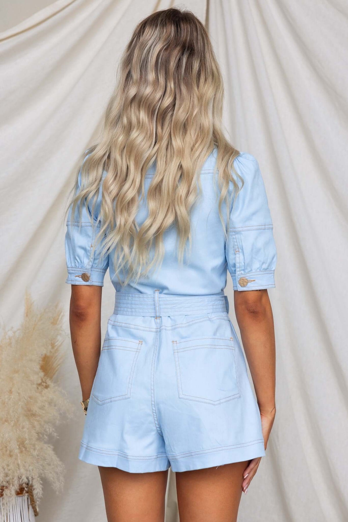 Back view of a woman wearing a light blue denim playsuit with elastic sleeves and waistband.