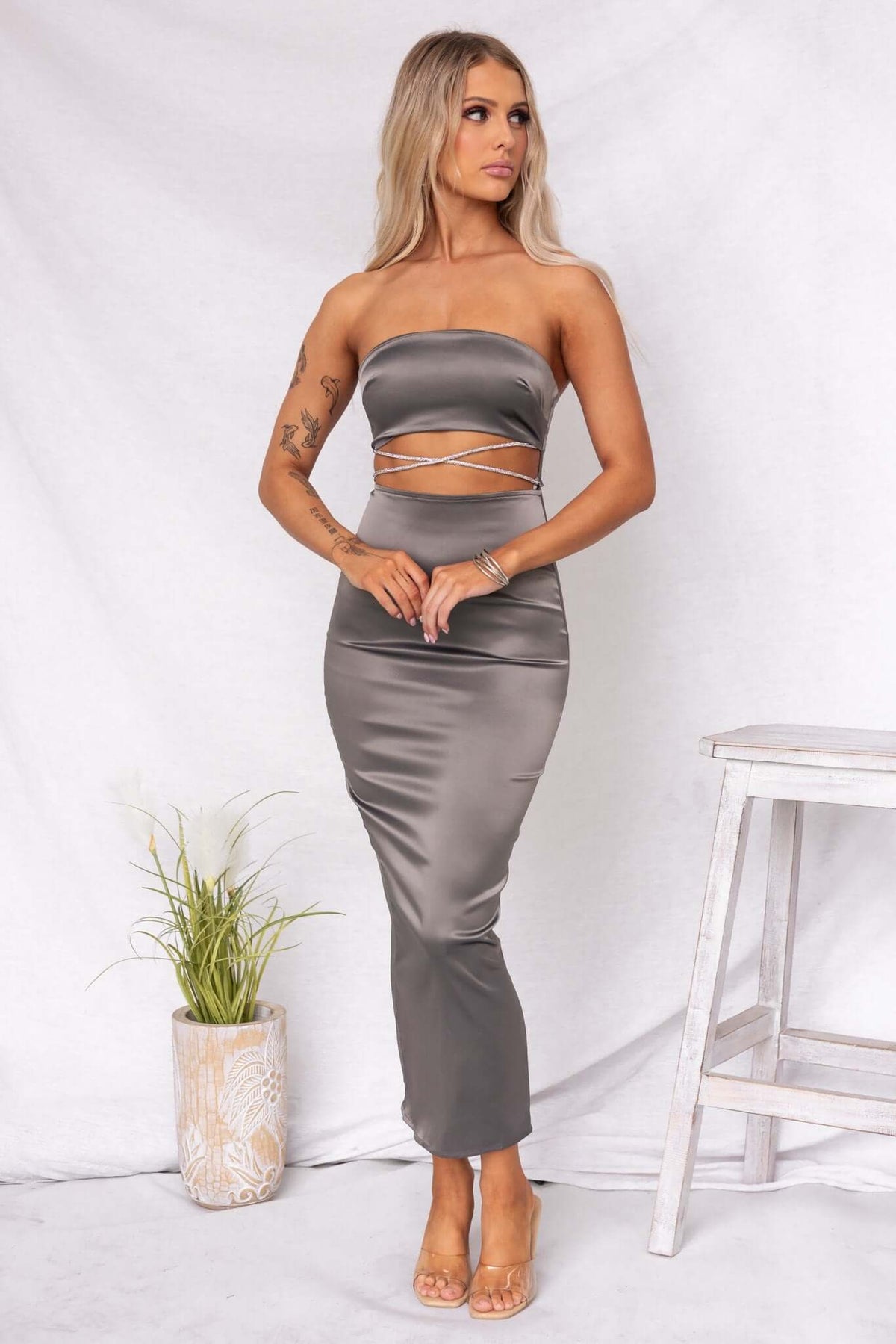 Satin strapless Louise Maxi Dress with cut-out detail and adjustable tie side, styled elegantly with a plant backdrop.