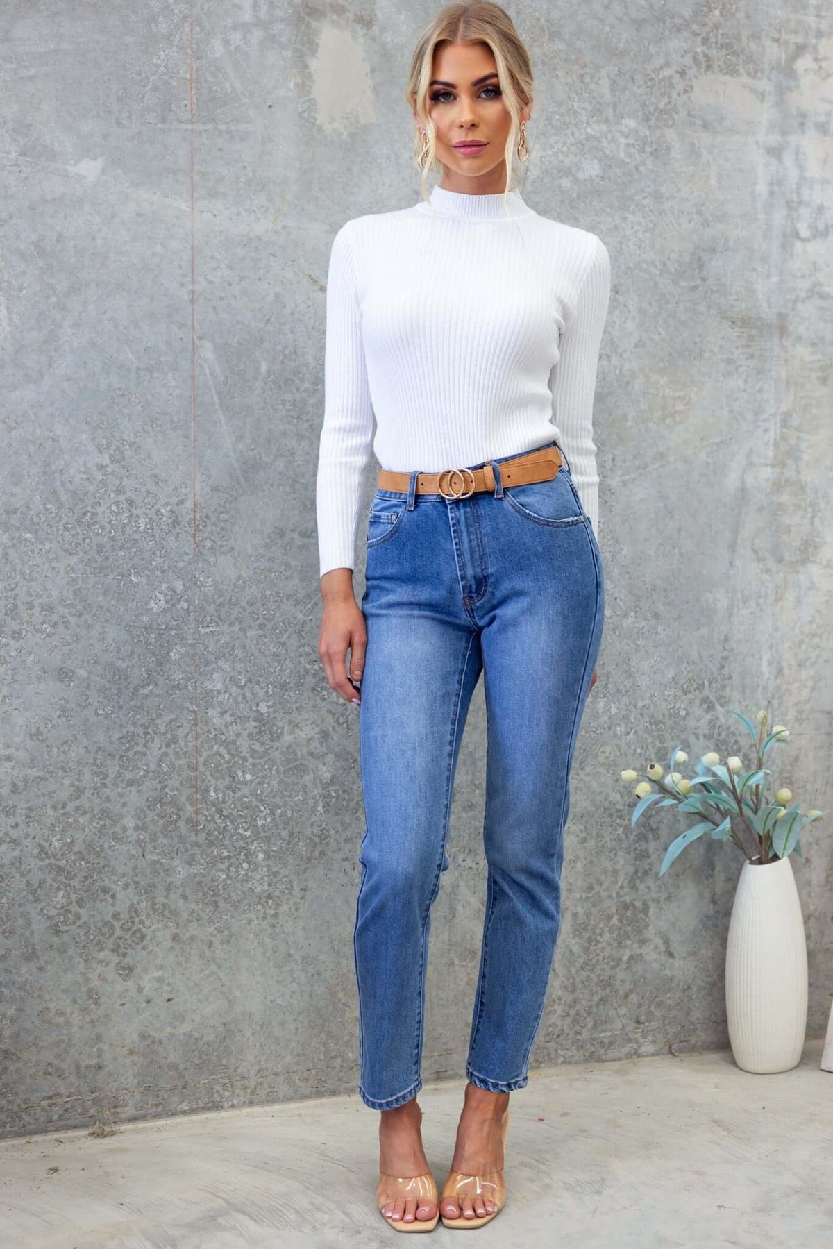 Model wearing Selma Jeans with mid-high waist, straight leg fit, paired with a white top and stylish belt, standing against a textured backdrop.