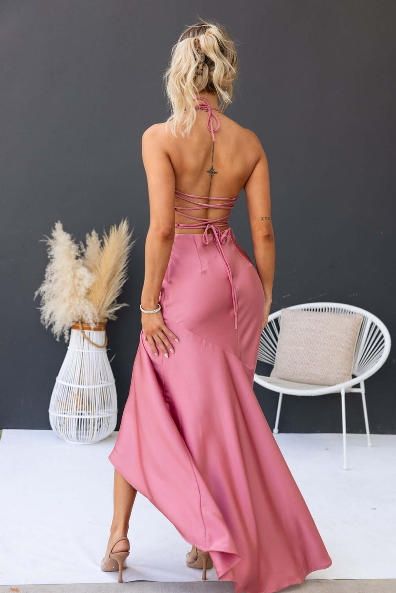 Back view of a woman in a pink Ramona maxi dress with tie neckline and flowing skirt, set against a neutral background.