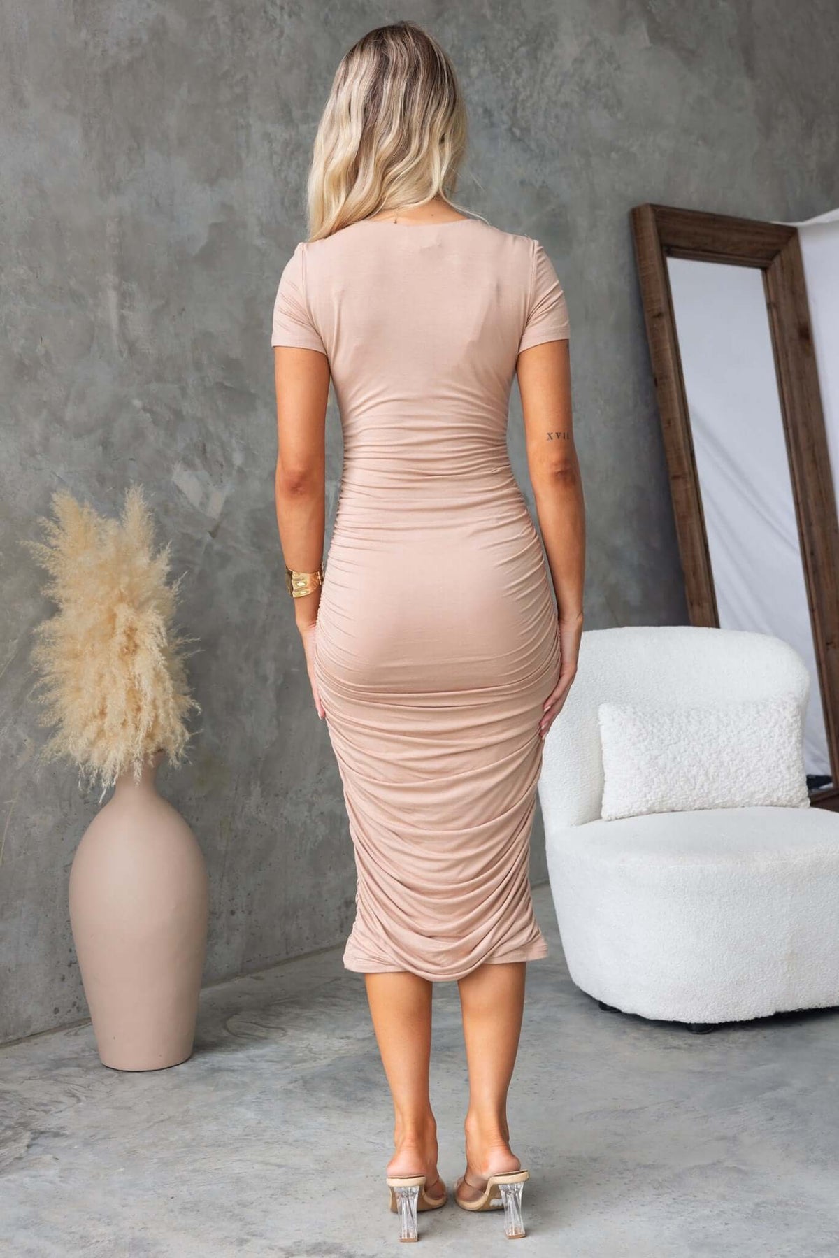 Back view of a woman in a beige bodycon midi dress with ruching, styled on a modern background.