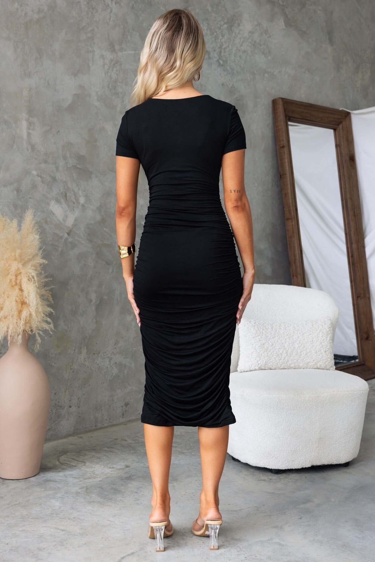 Back view of a woman in a black bodycon midi dress with side ruching, styled against a modern minimalist backdrop.
