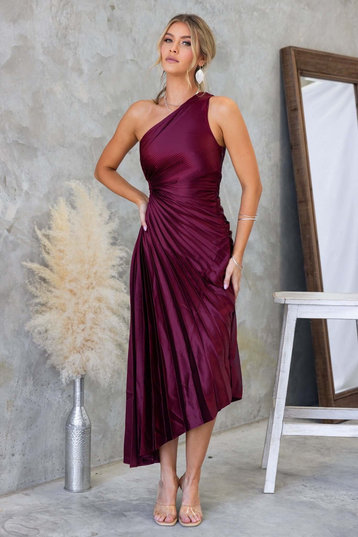 Model wearing a burgundy one-shoulder maxi dress with pleats and cut-out waist, styled in a modern room.