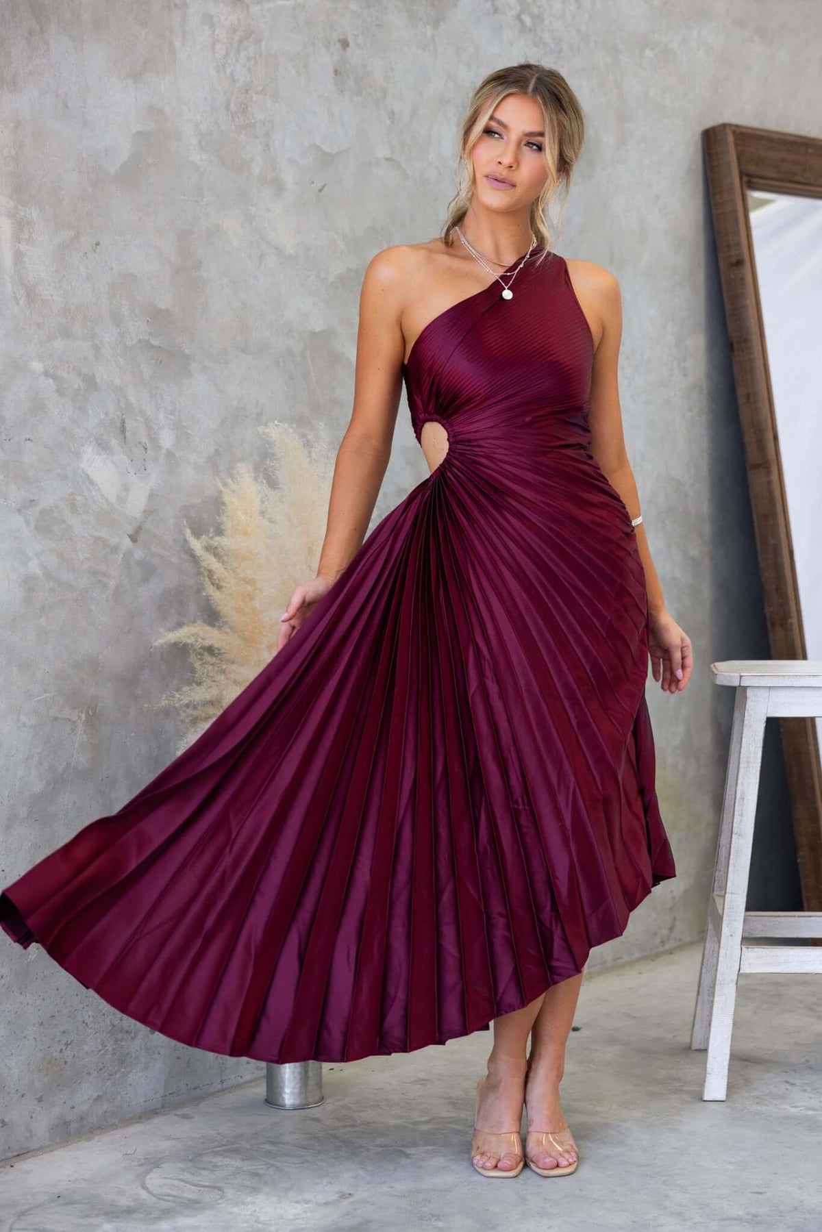 Woman wearing a burgundy Olympia Maxi Dress with one-shoulder design and pleated skirt in a stylish setting.