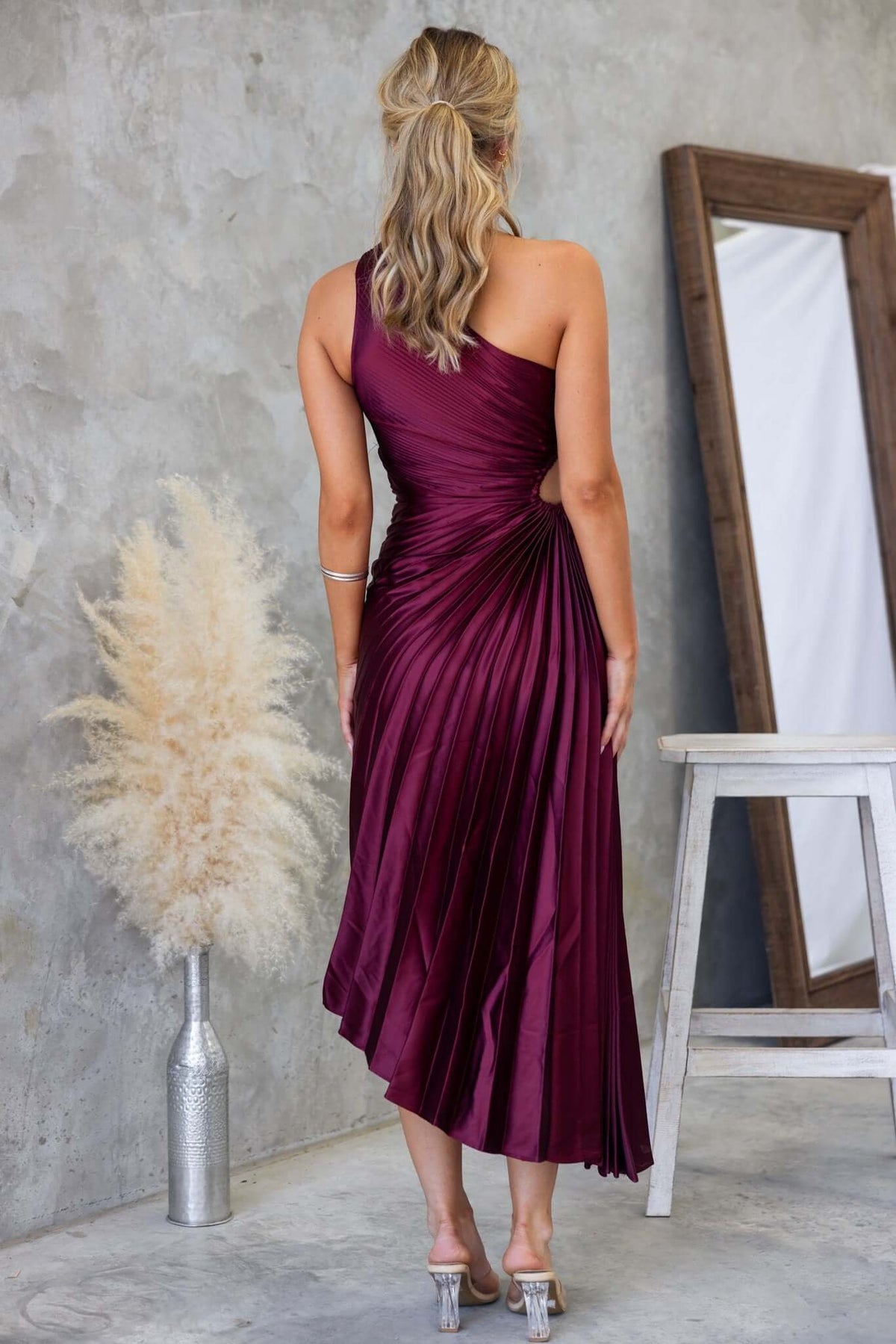 Back view of a woman in a burgundy Olympia maxi dress with a one-shoulder neckline and accordion pleats.