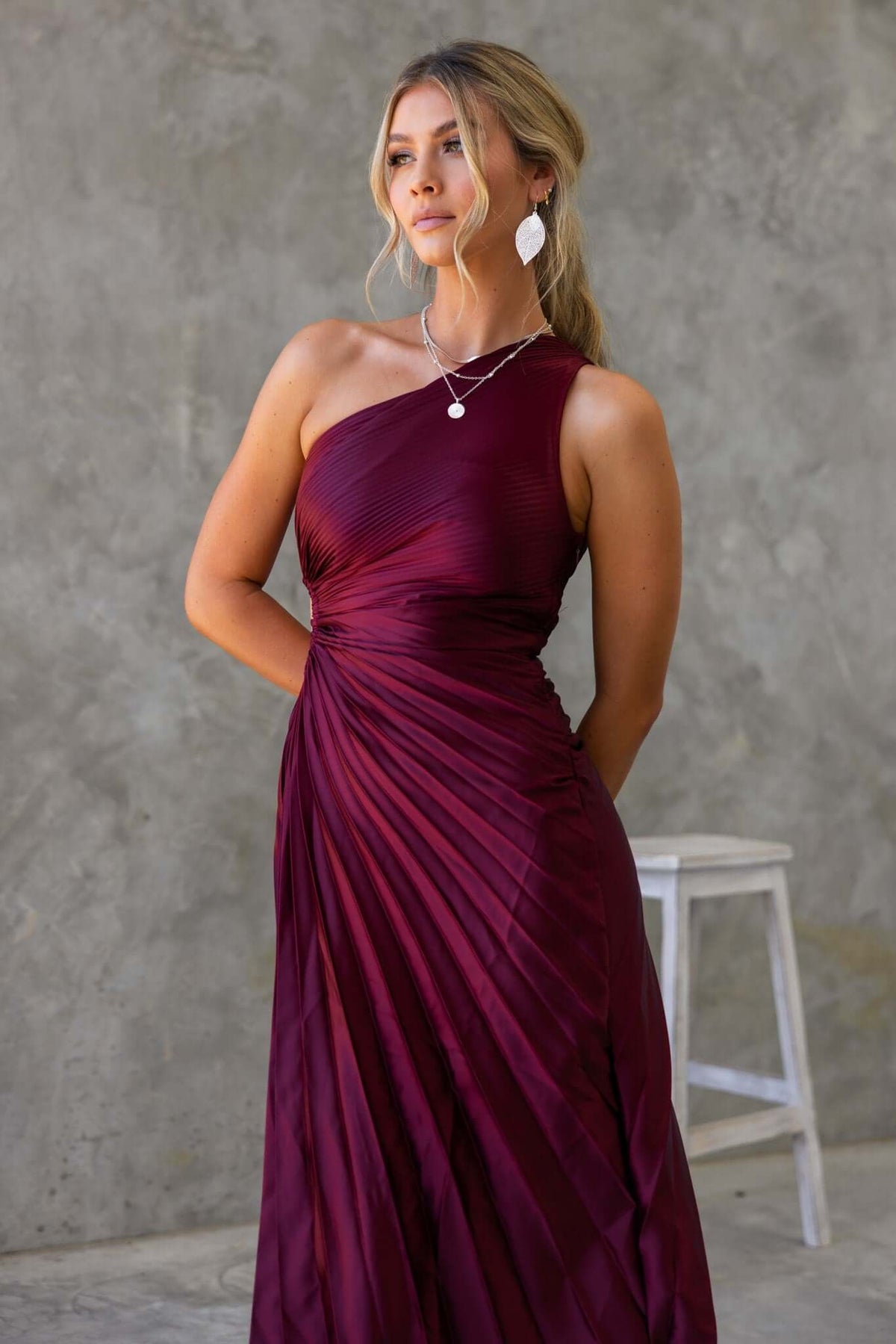 Model wearing a burgundy one-shoulder Olympia Maxi Dress with accordion pleats and a cut-out waist.