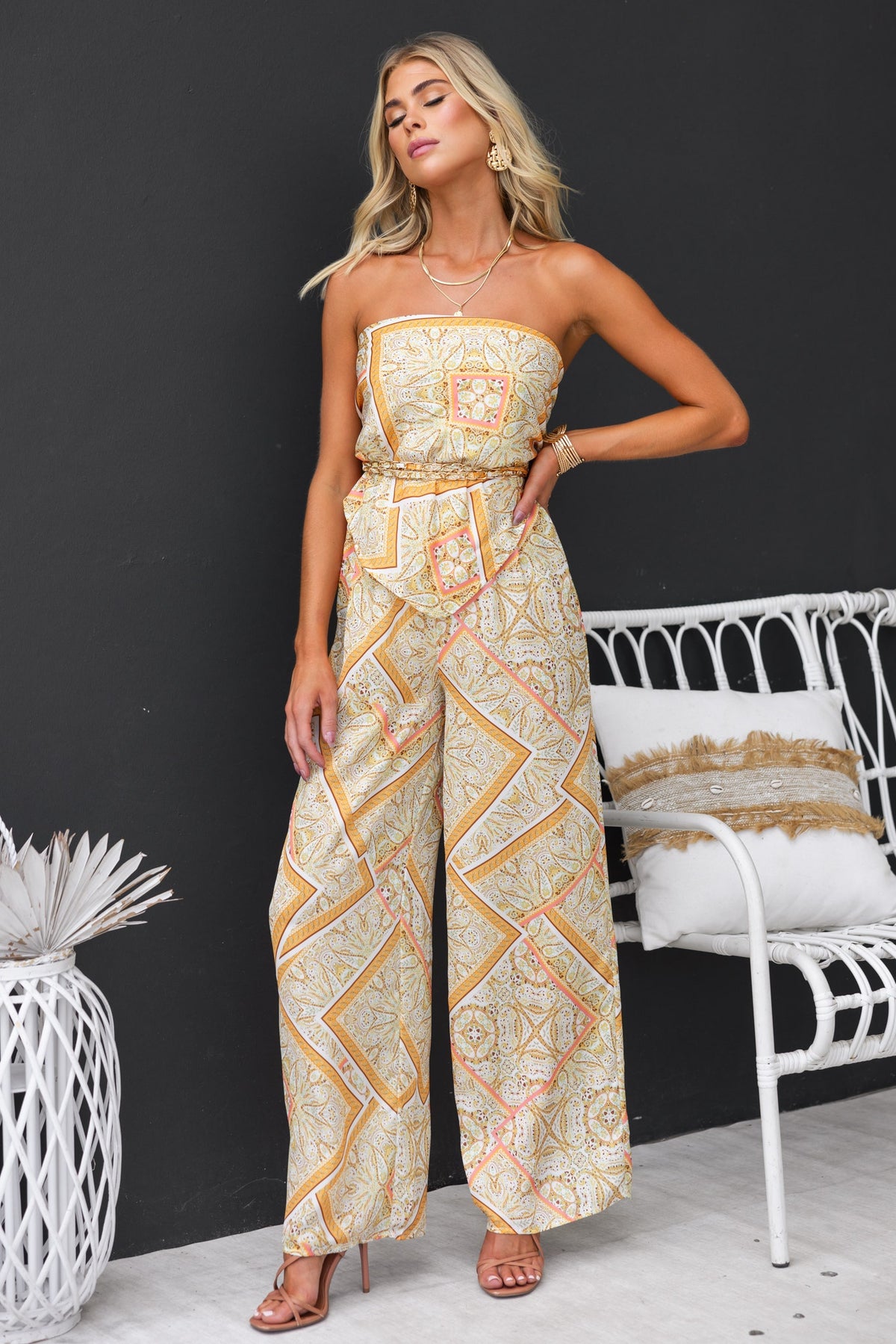 Woman in abstract print strapless jumpsuit with wide legs, styled with sandals, against a modern backdrop.
