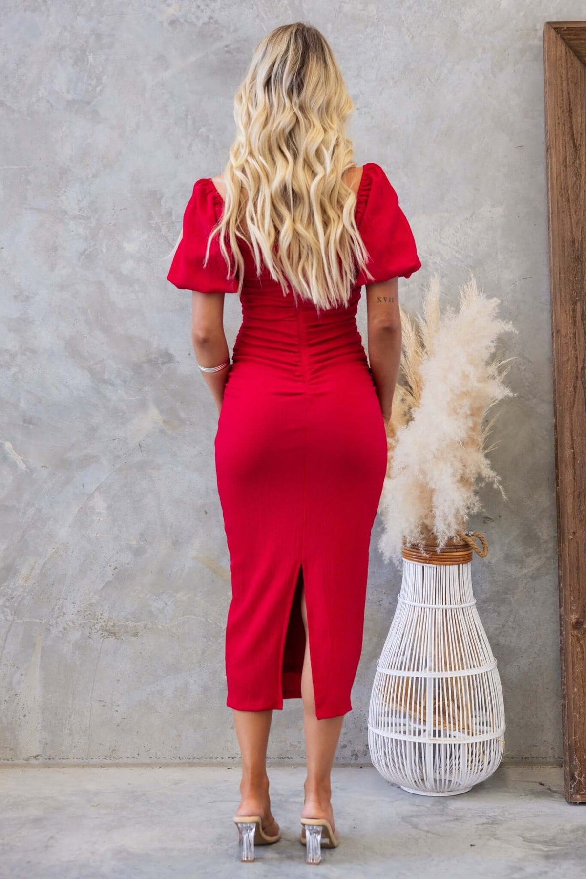 Back view of Topeka Midi Dress, featuring puff sleeves, ruching at sides, and a back split, styled with a plant backdrop.