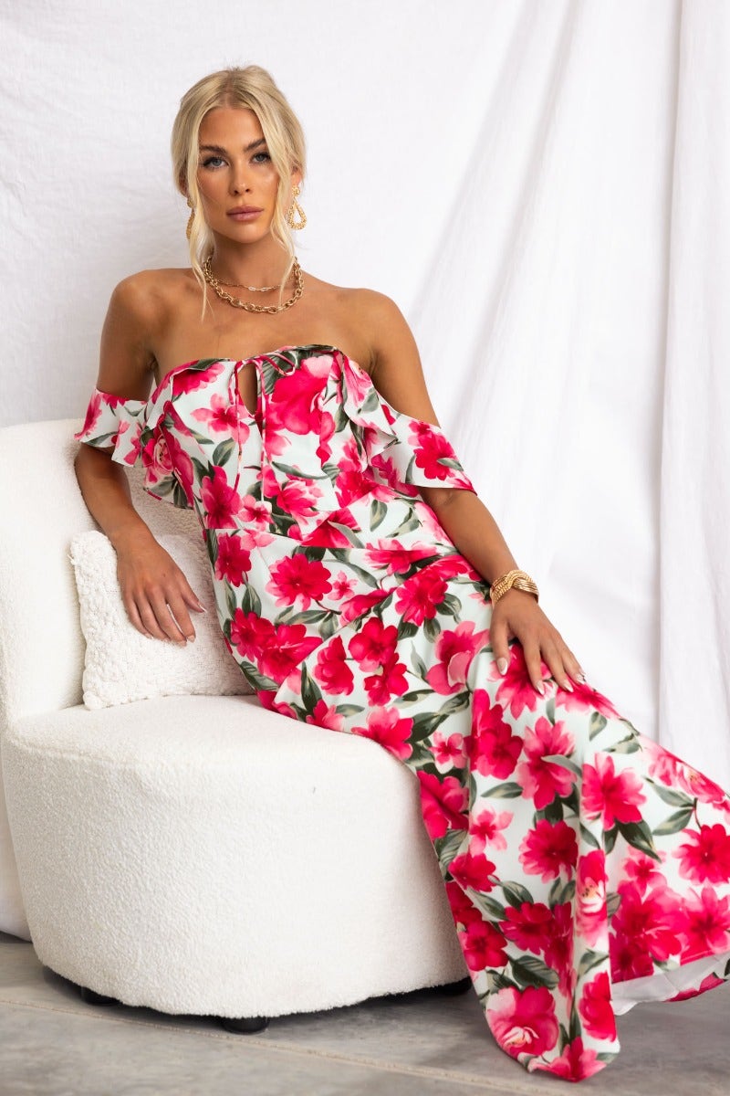 Model wearing a floral print off-shoulder maxi dress with tie detail and side zip, seated on a lounge chair.