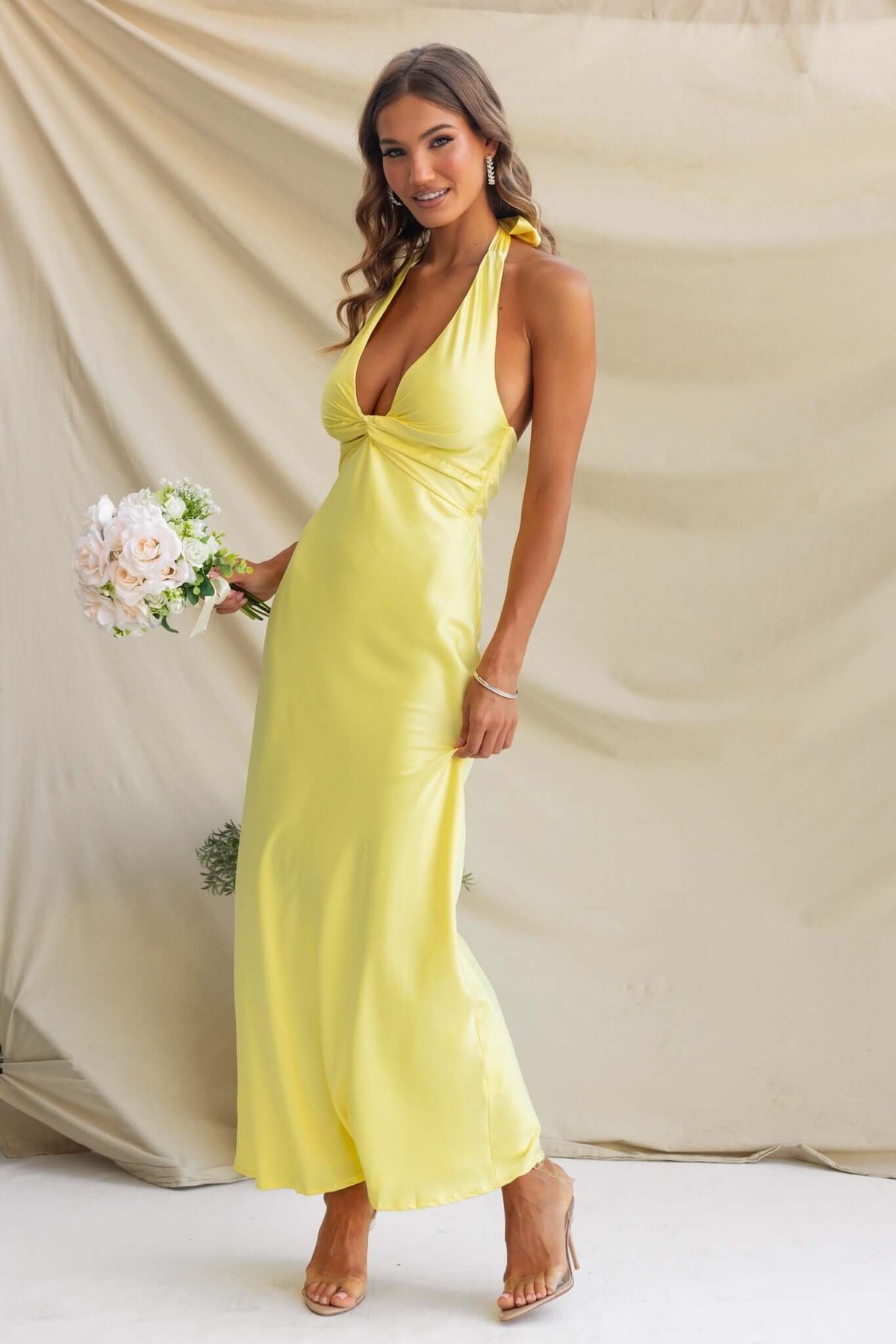 Model wearing a yellow satin tulip maxi dress with a V-neckline, holding flowers against a beige backdrop.