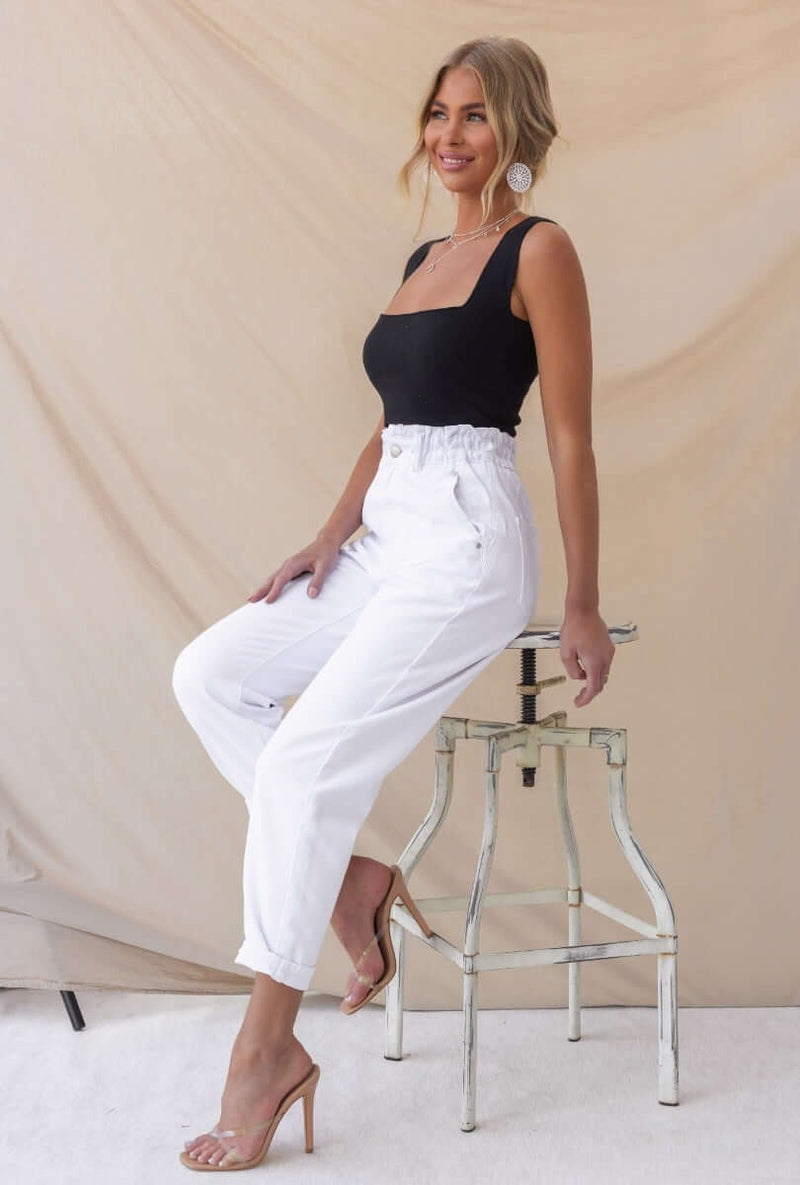 Woman wearing Cameron Paperbag Jeans with white fabric and a black top, styled sitting on a clear stool.
