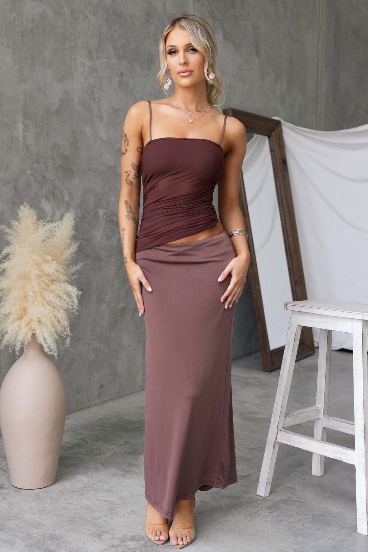 Woman wearing a brown Denham maxi dress with adjustable straps and a fitted silhouette, styled in a modern indoor setting.
