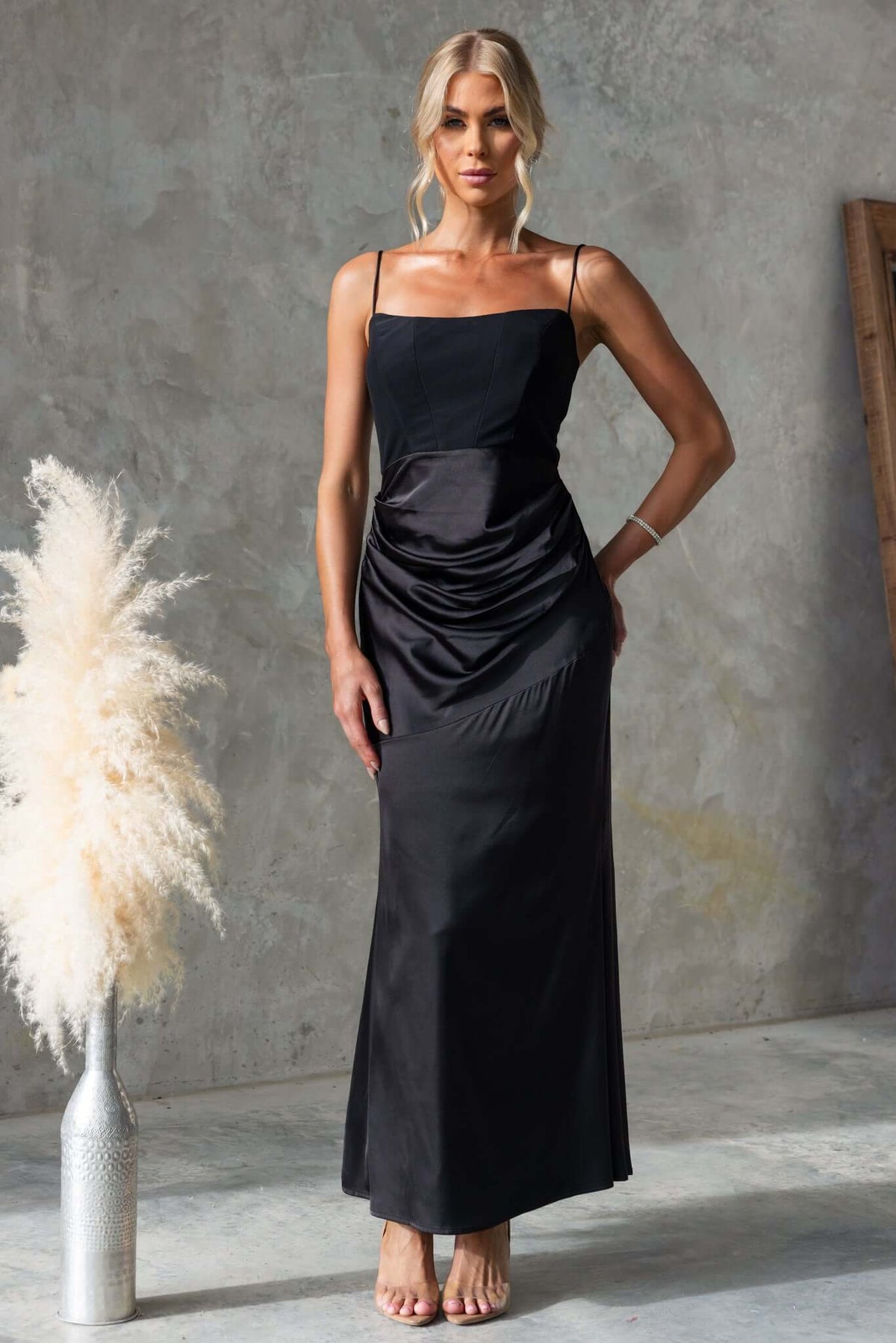 Amaroso Bridal Atheni Maxi Dress in black with adjustable straps and satin skirt, stylishly posed against a minimalist backdrop.