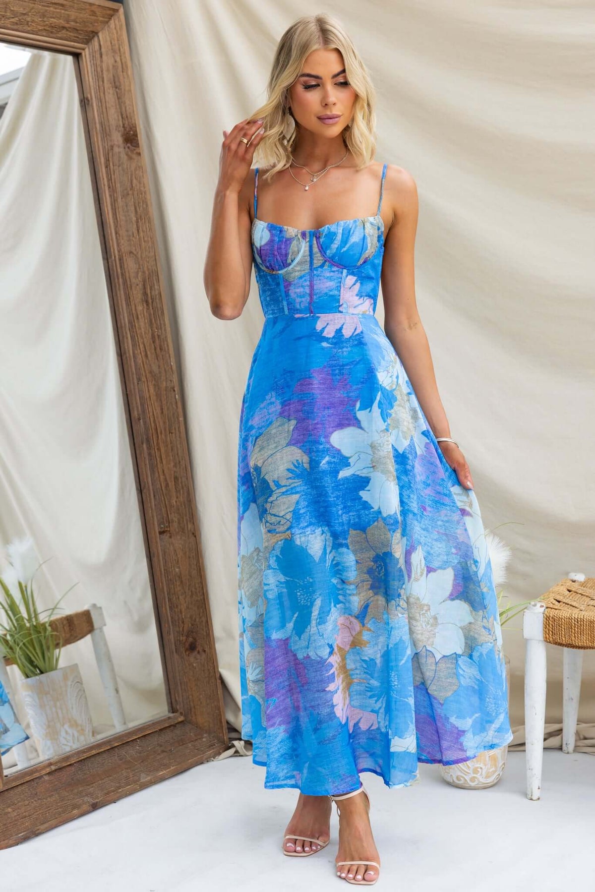 Model wearing a floral Burkes Midi Dress in blue, featuring adjustable straps and a fitted bodice, styled in a bright setting.