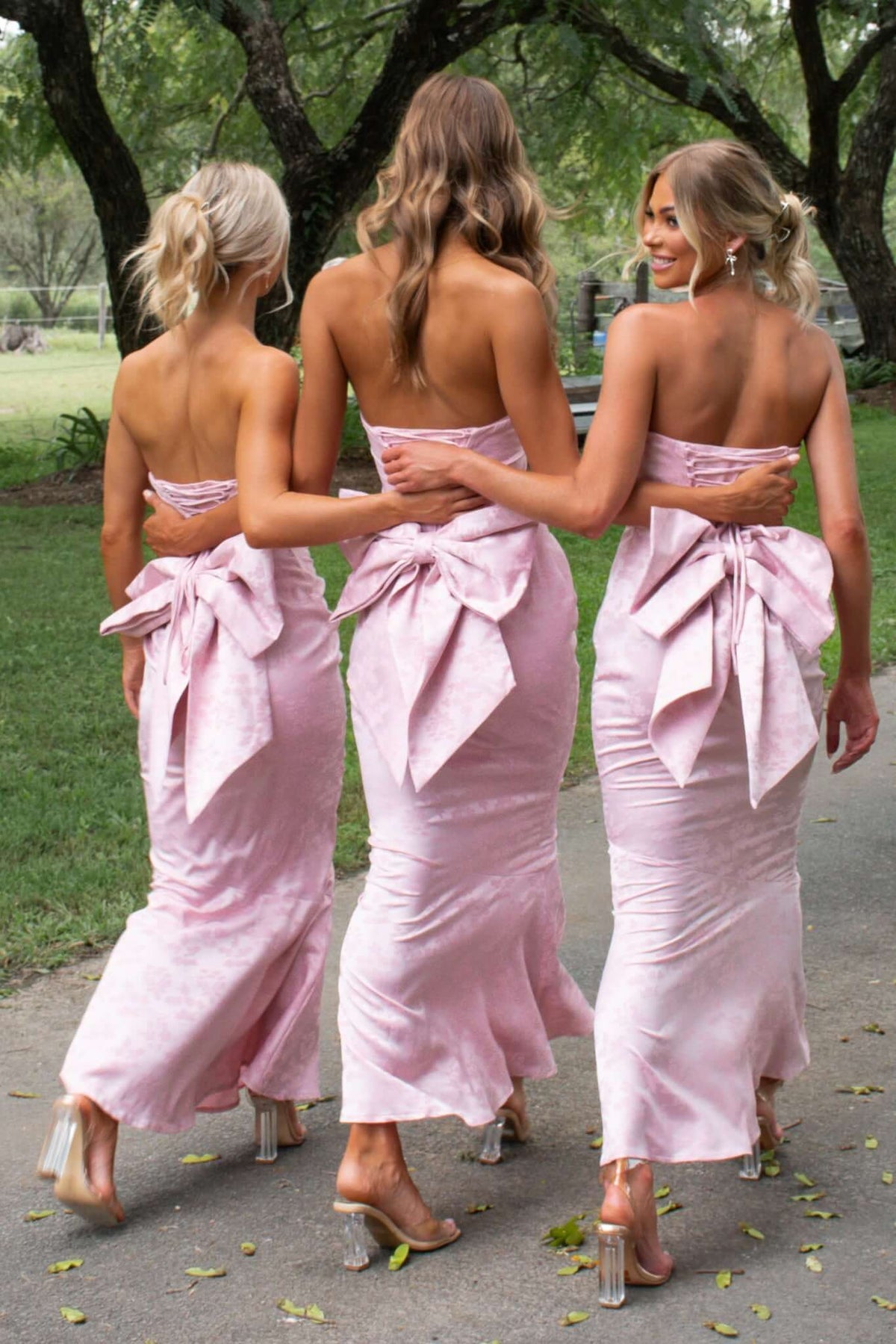 Three women in pink strapless maxi dresses with large bows at the back, walking in a scenic outdoor setting.