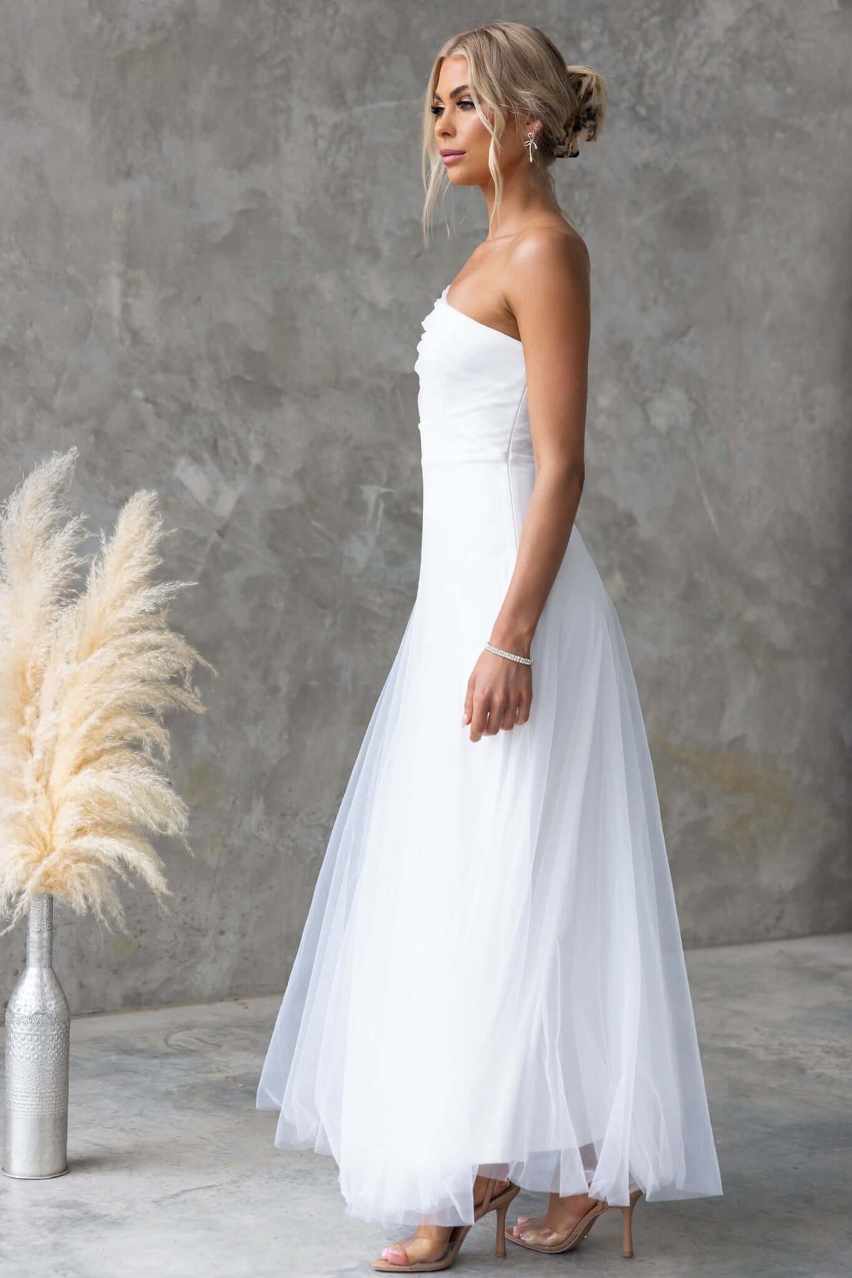 Eleah Maxi Dress in white, featuring a one-shoulder neckline and tulle material, styled elegantly against a textured background.