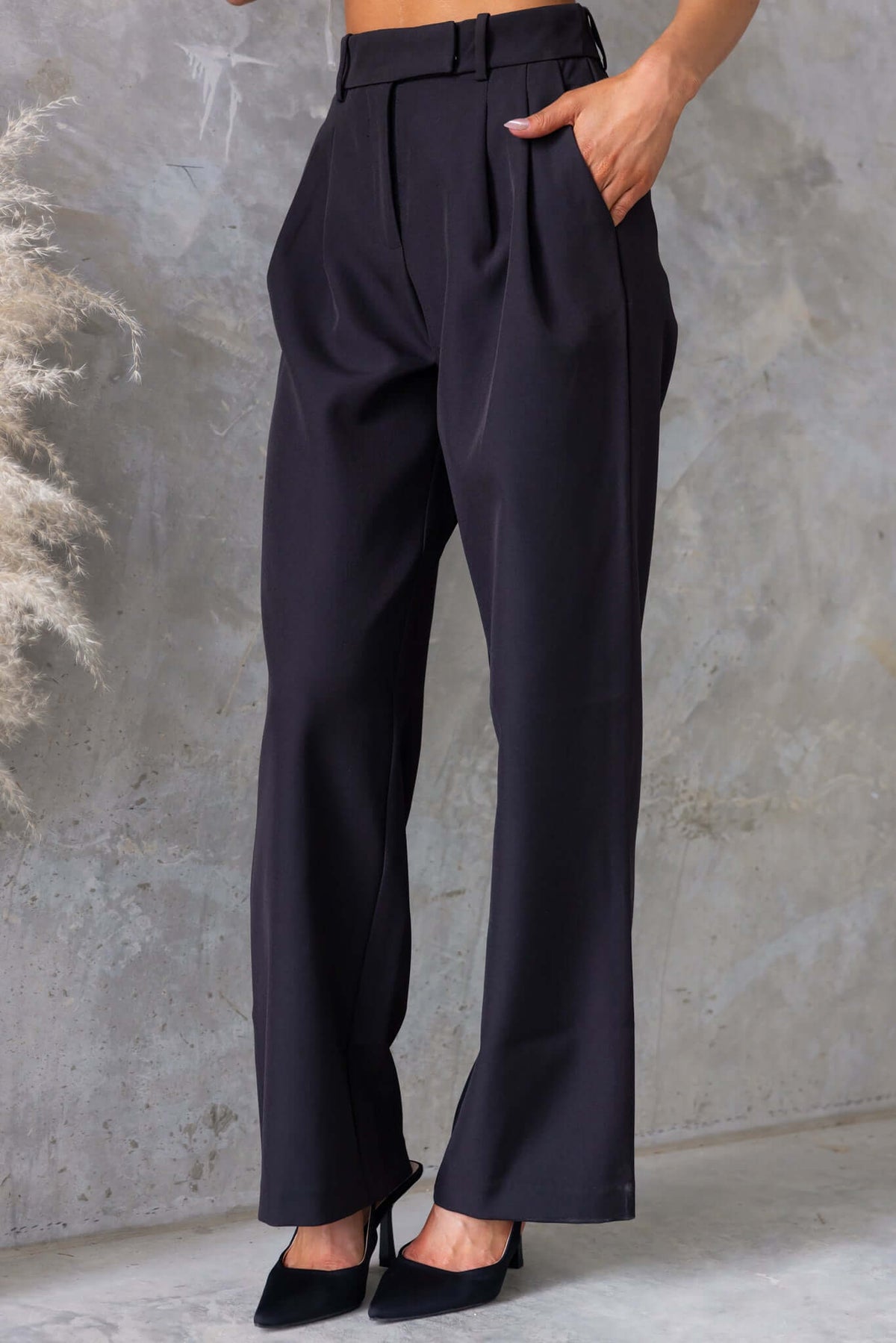 Gette Pants in black with mid-high waist, pockets, and a tailored fit, styled with heels against a textured backdrop.