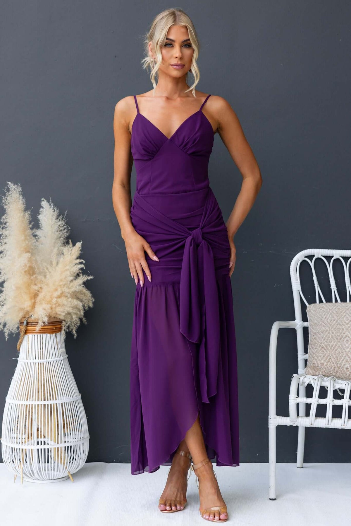 Model wearing a purple Iconic Maxi Dress with adjustable straps and front split, styled with a chic backdrop.