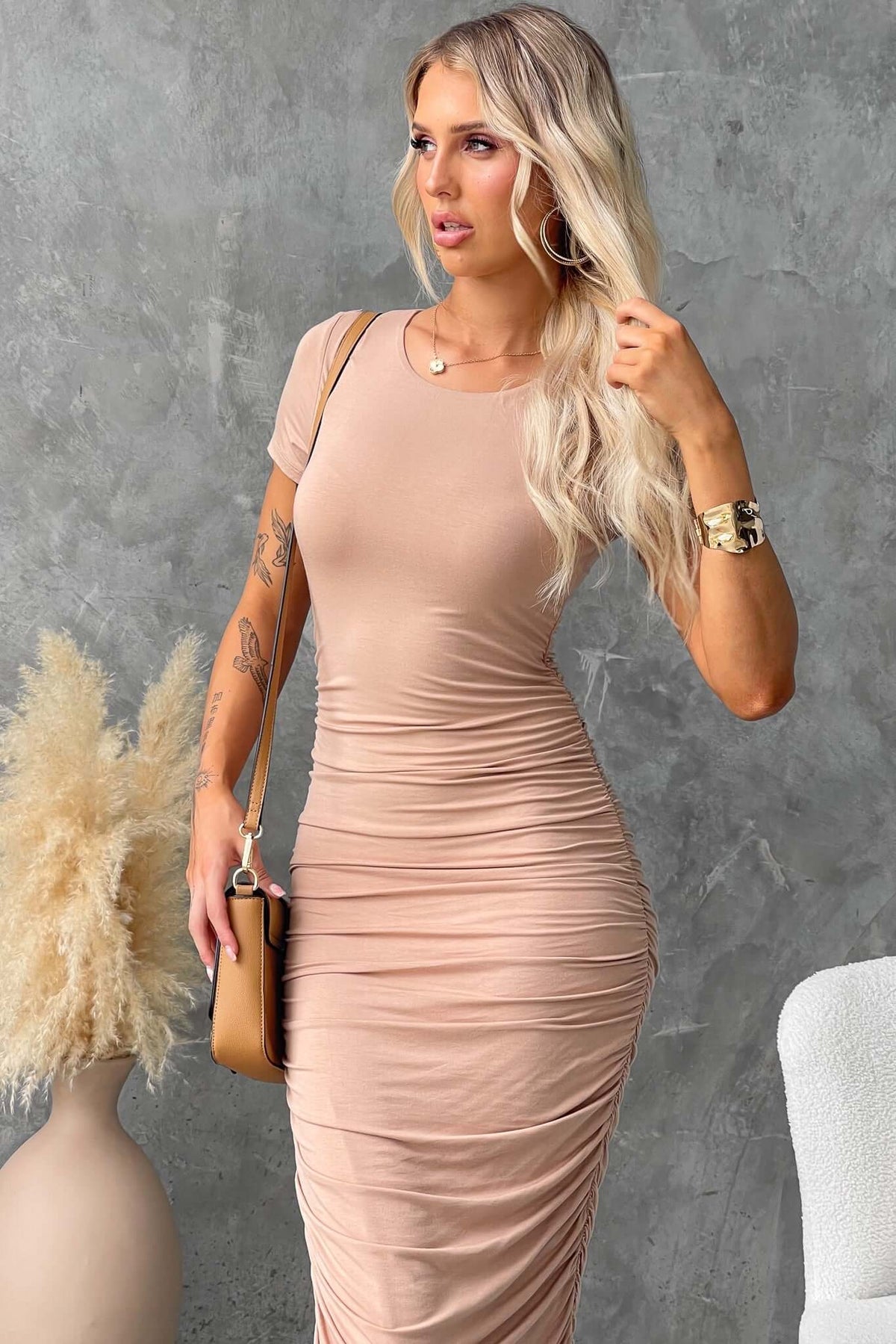Model wearing a bodycon midi dress with side ruching, styled against a textured backdrop and a decorative vase.
