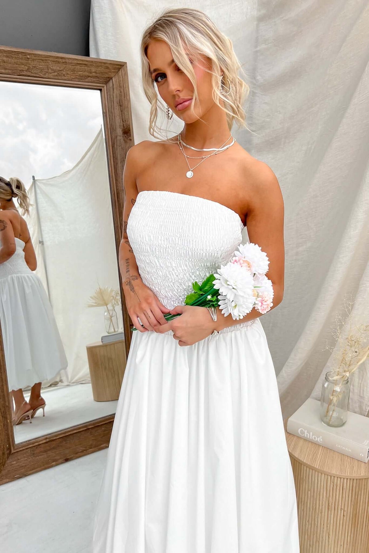 Model wearing a strapless white Cadro Midi Dress, holding flowers, with a mirror in background.