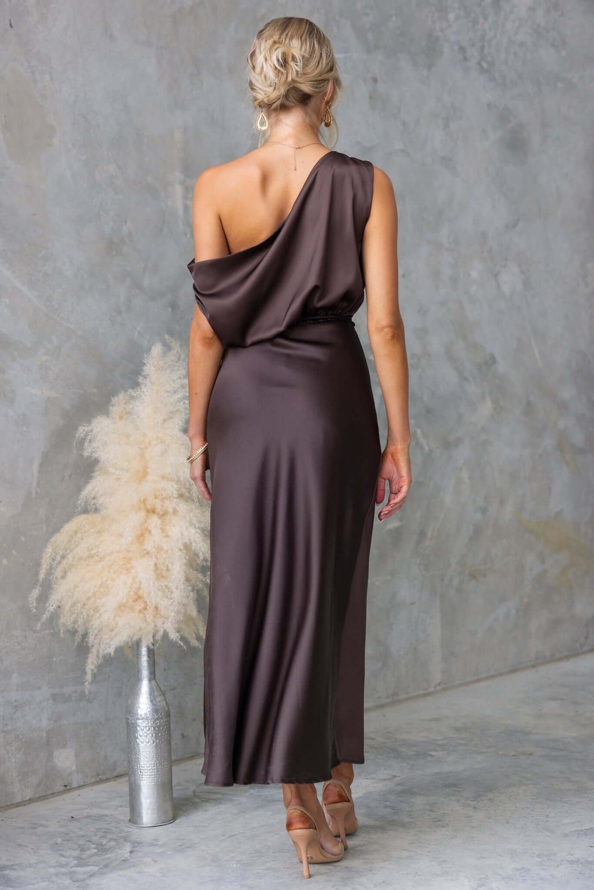 Back view of Issey Maxi Dress, featuring asymmetric neckline and satin fabric, styled with pumps and decorative plant.