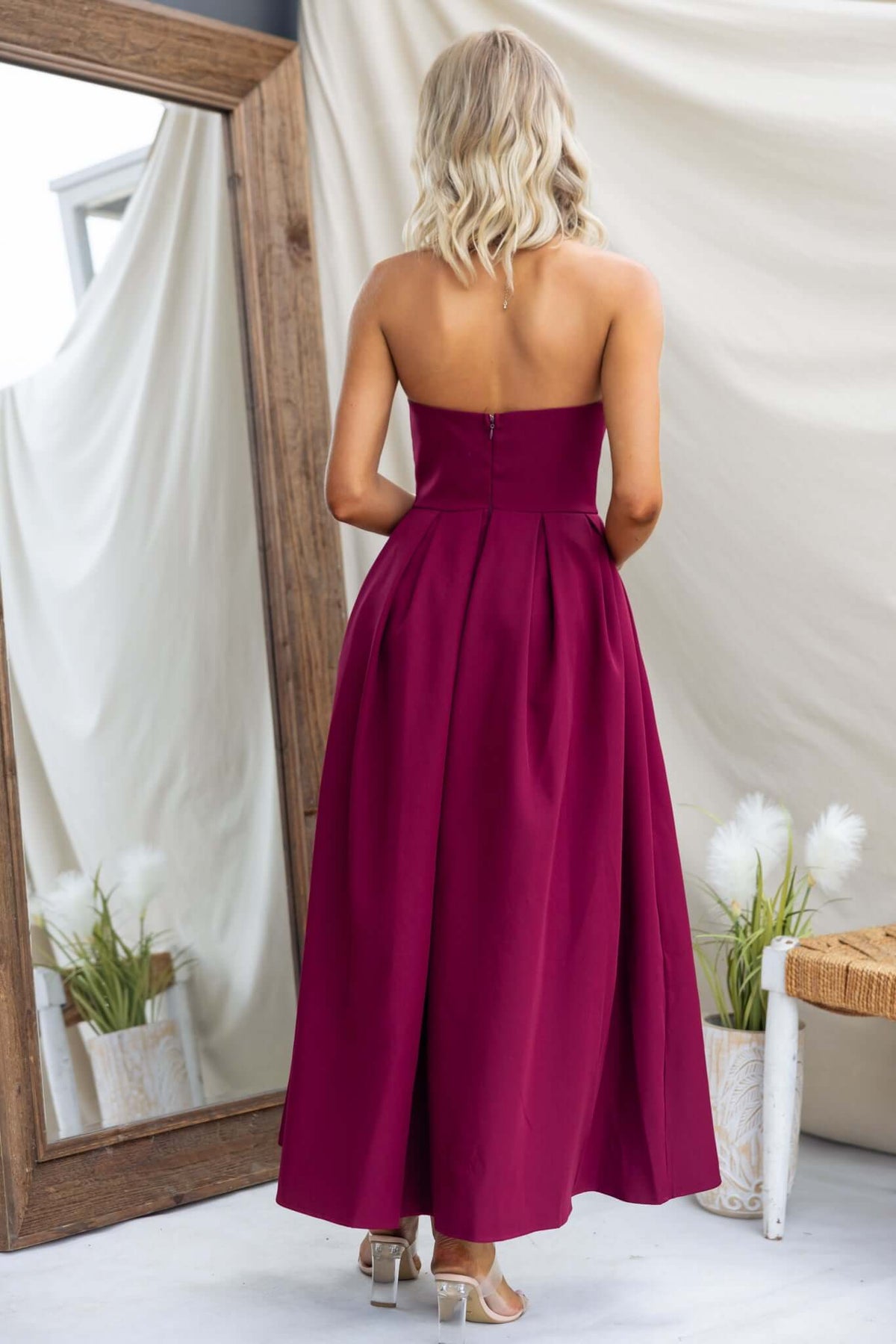 Back view of a woman in a burgundy strapless maxi dress with pleats, showcasing an elegant silhouette.